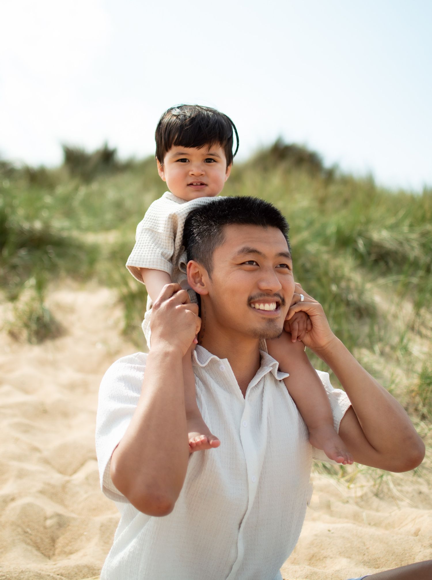 Smiling father carries his young toddler on his shoulders on a sunny sandy beach.