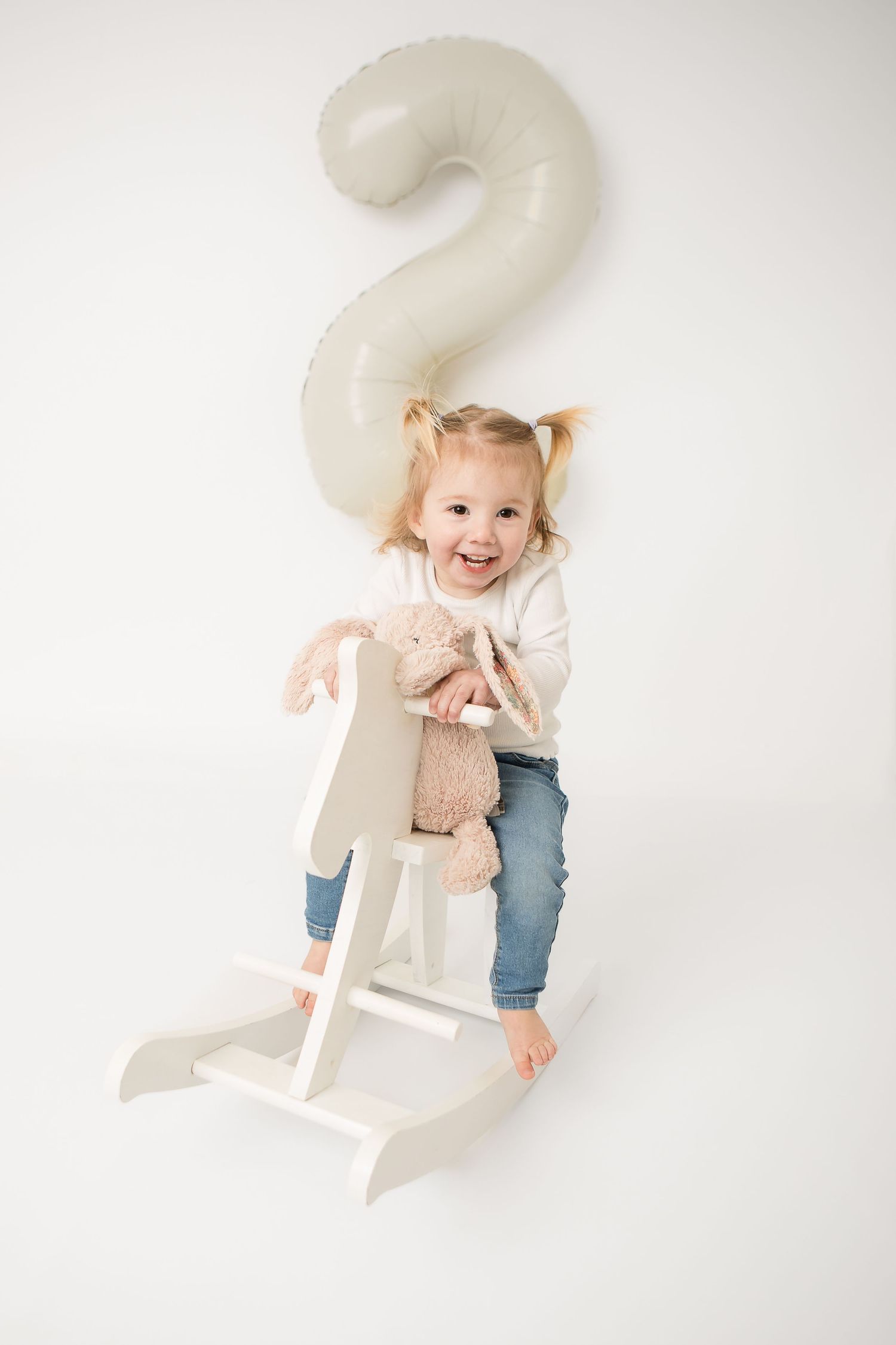 Smiling toddler girl riding a white rocking horse holding a stuffed bunny with a number 2 balloon behind her.