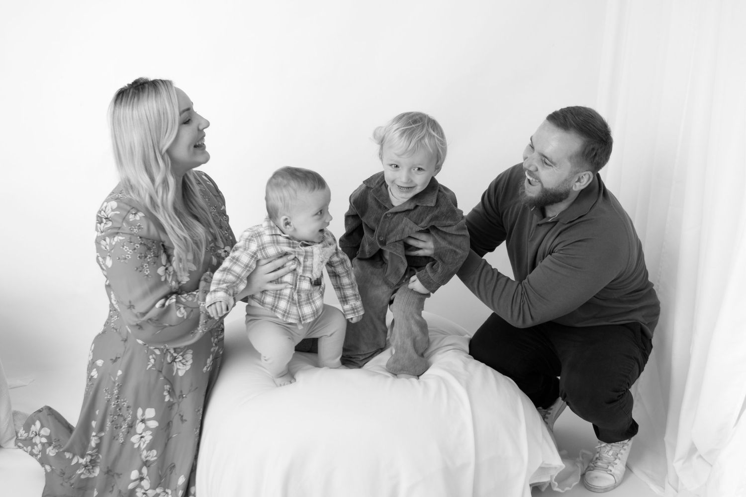Black and white family photo of parents with two young boys laughing and playing together in a bright studio setting.