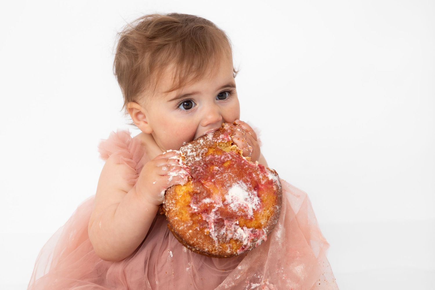 Adorable baby girl in pink tutu eating a large glazed donut covered in powdered sugar on white background.