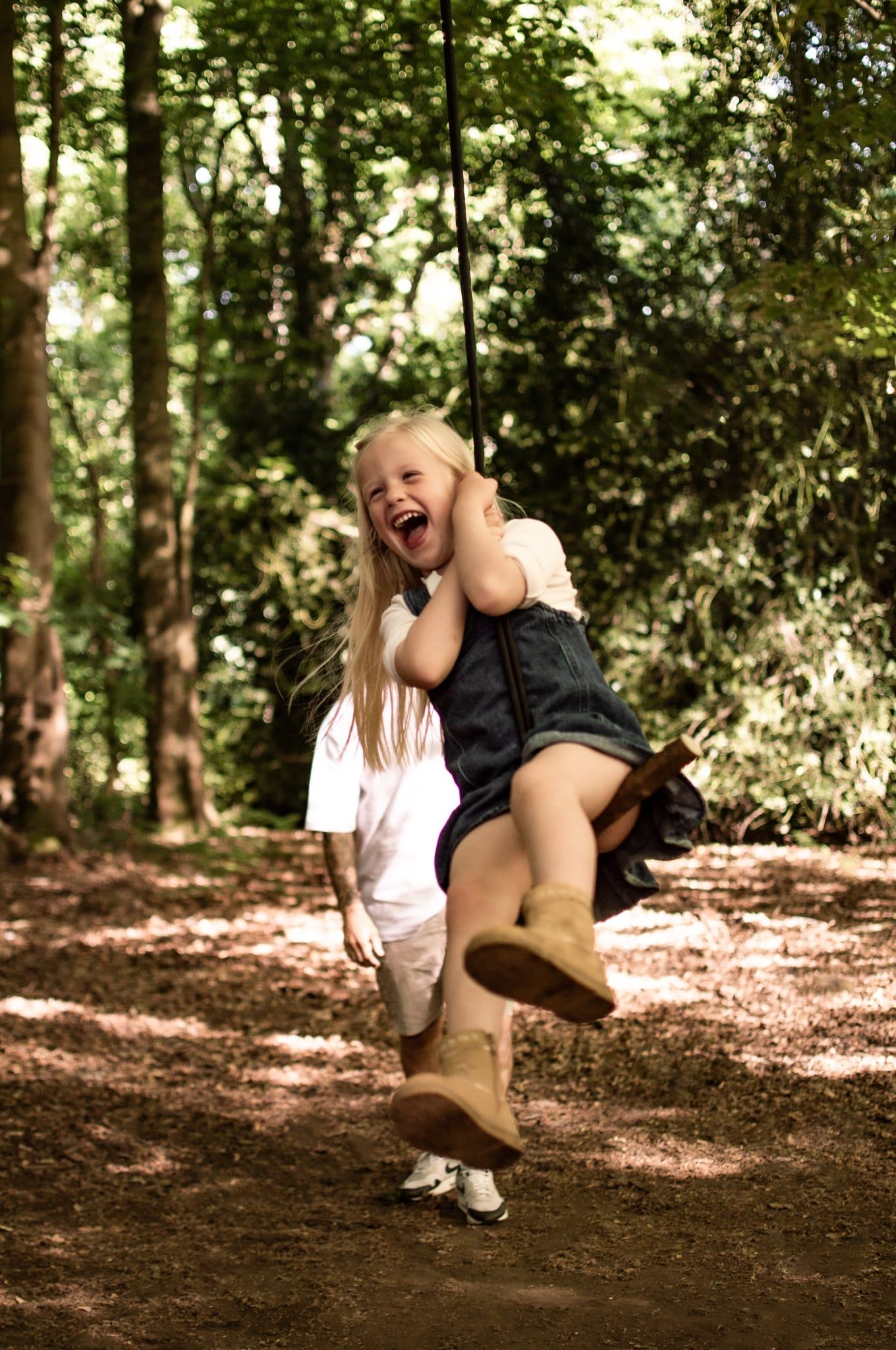 Two young girls laughing and playing on a rope swing in a sunlit forest, one riding while the other pushes.