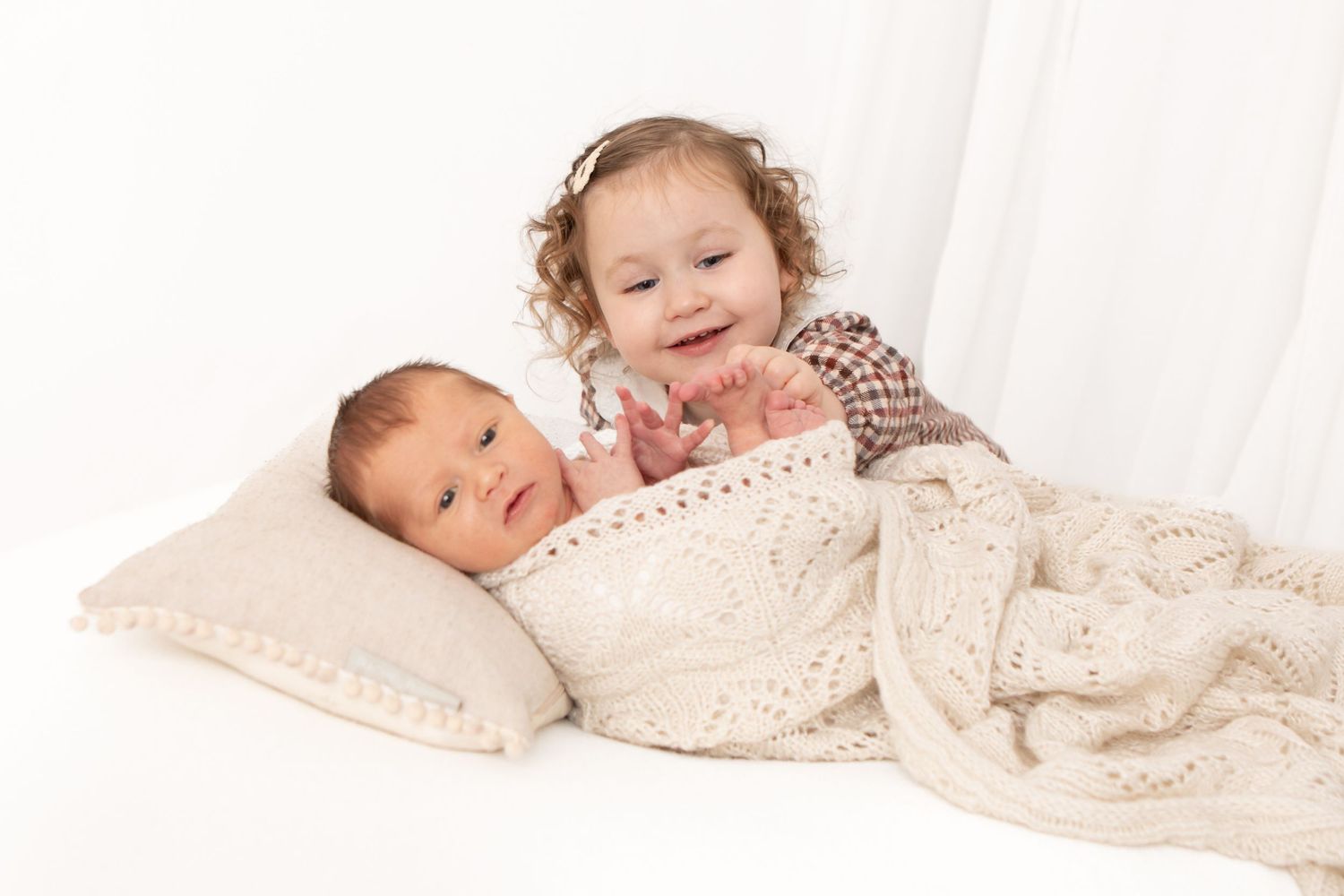 Siblings resting together under delicate lace blanket in soft natural light.