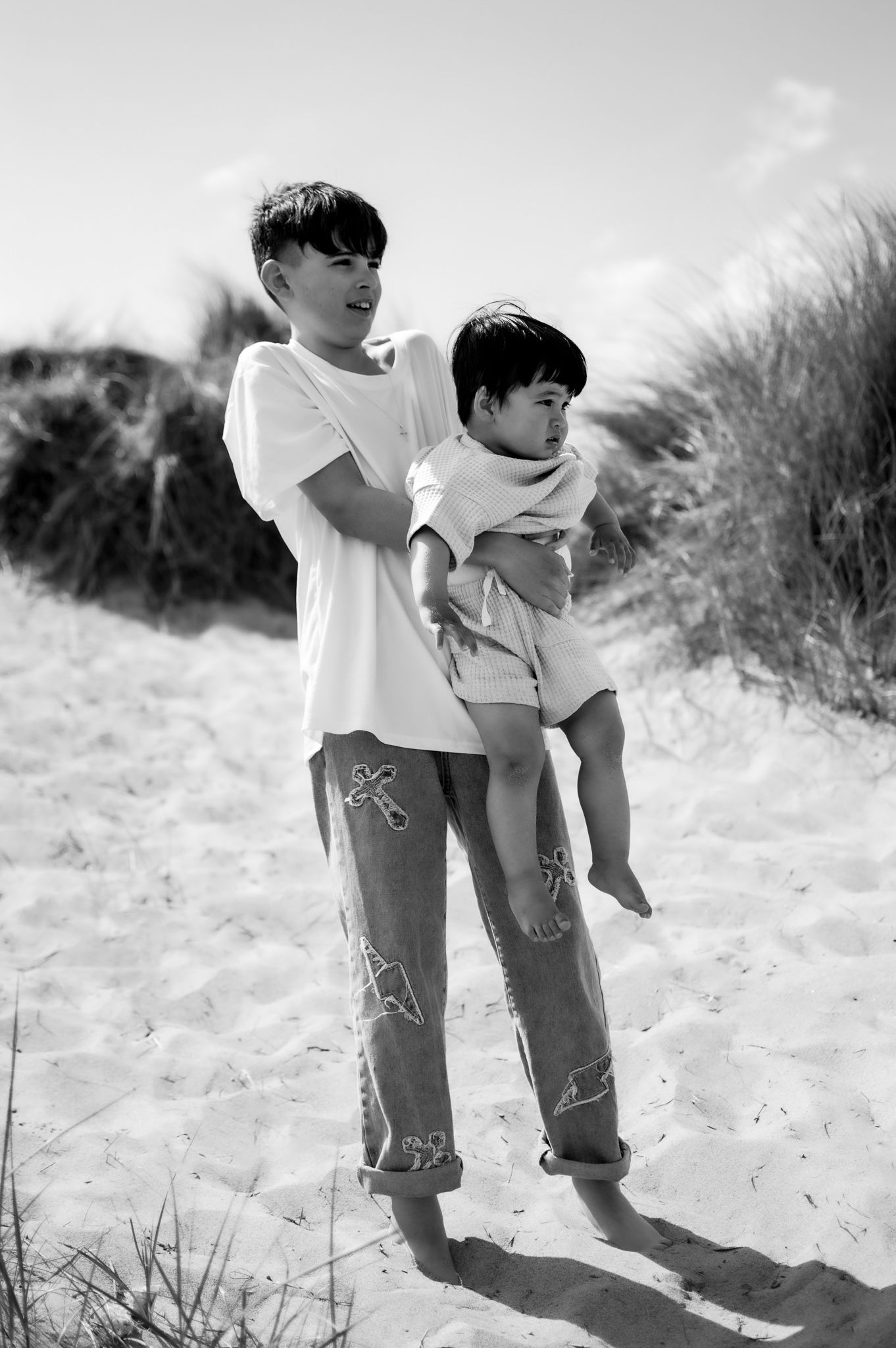Black and white photo of an older boy carrying a younger child on a sandy beach with dunes in the background.