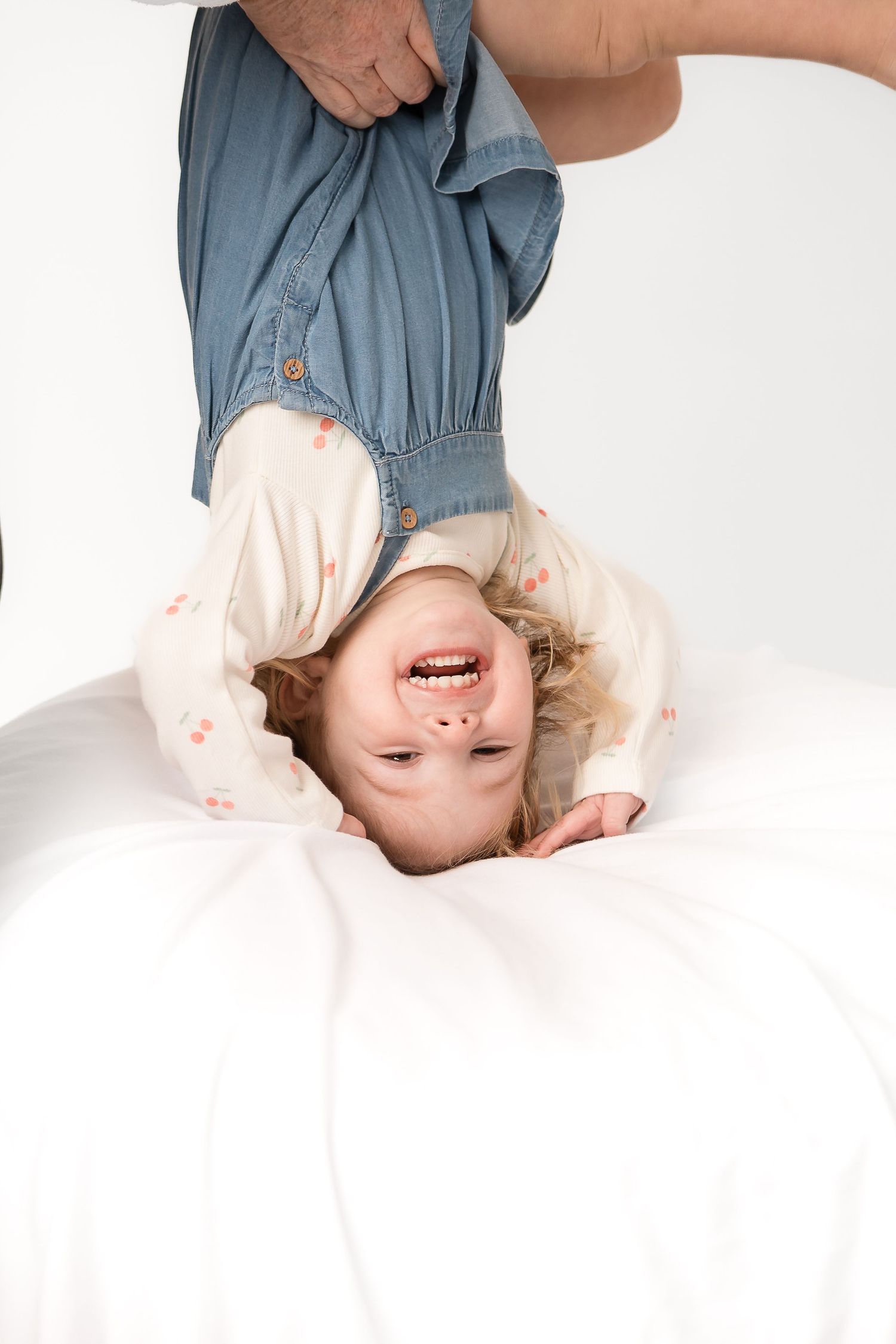 Joyful toddler held upside down by adult hands, laughing with arms outstretched over white surface.