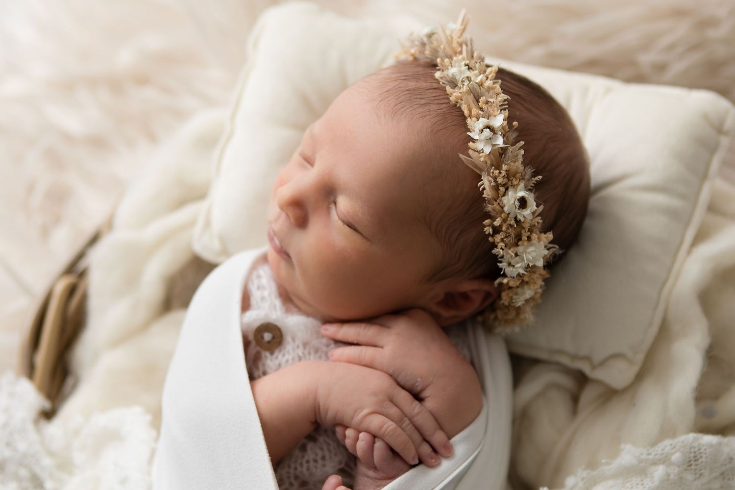 Newborn baby wearing delicate gold floral crown sleeps peacefully on cream colored bedding.