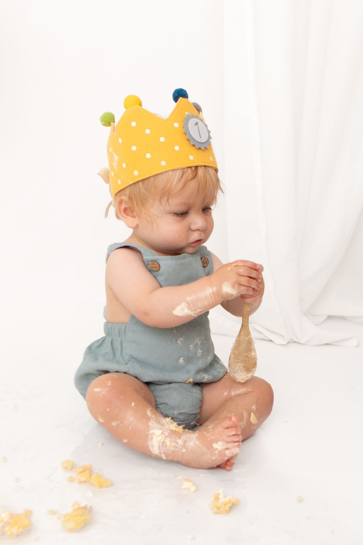 Baby in yellow crown hat and gray romper smashing first birthday cake, sitting on white background.