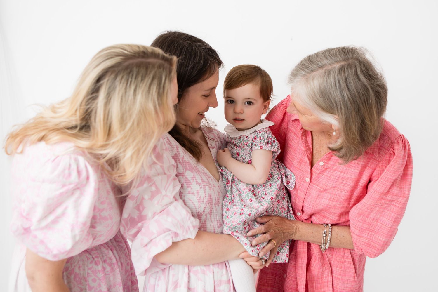 Three generations of women in pink outfits smiling at an adorable red-haired baby girl against a white background.