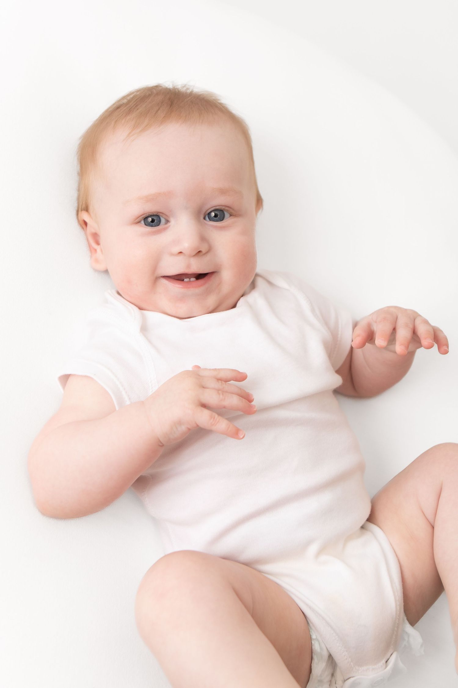 Smiling baby in white onesie lying on white background, looking at camera with blue eyes and rosy cheeks.