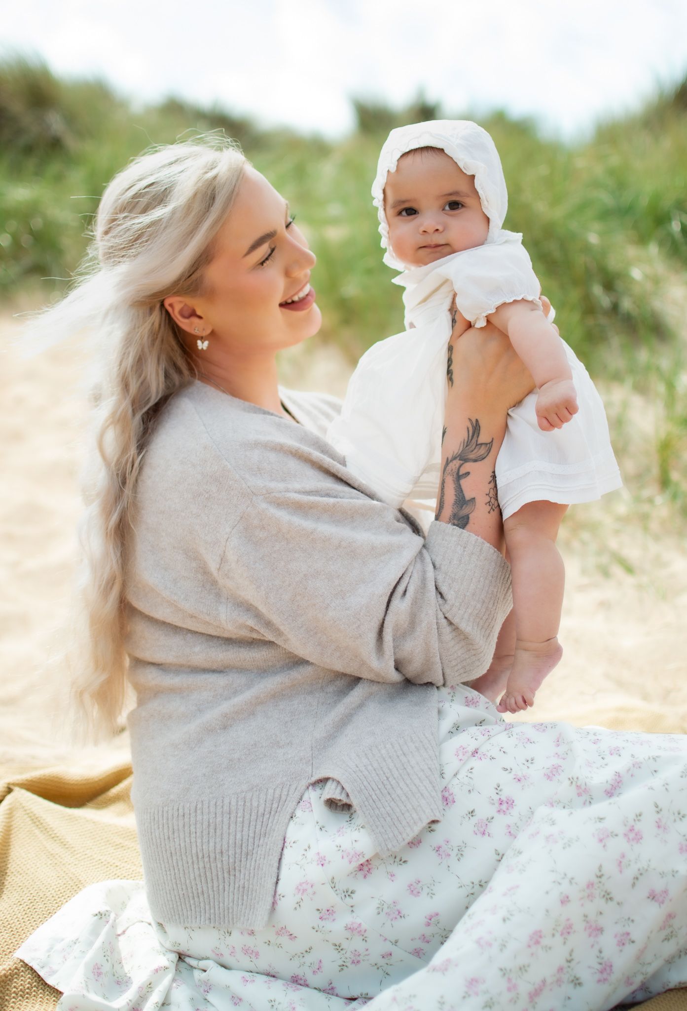 Blonde mother sitting on beach blanket holding smiling baby in white bonnet and dress among sandy dunes.