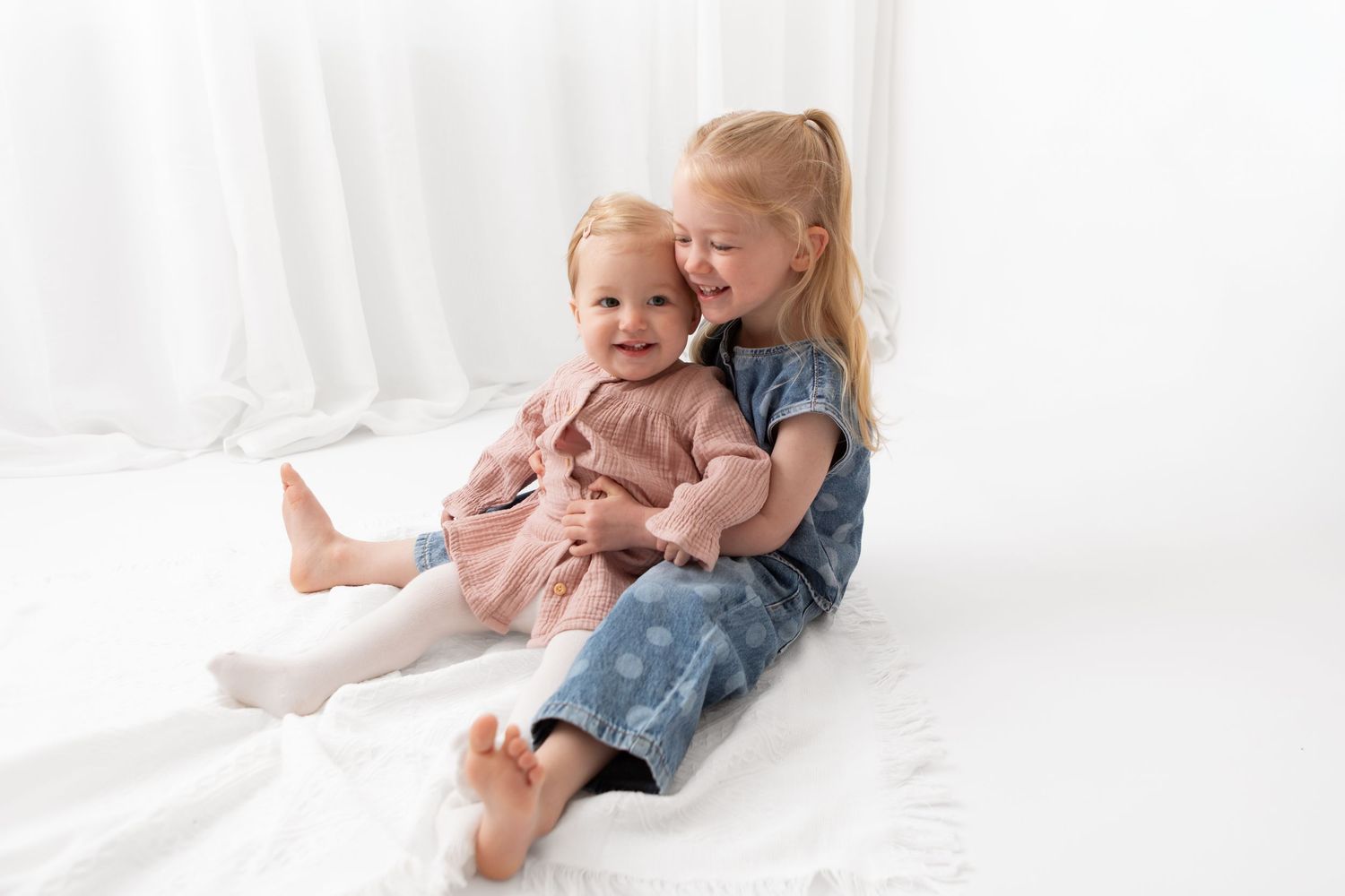Two young sisters in pink and blue dresses sitting together on white blanket, older girl hugging baby sibling.