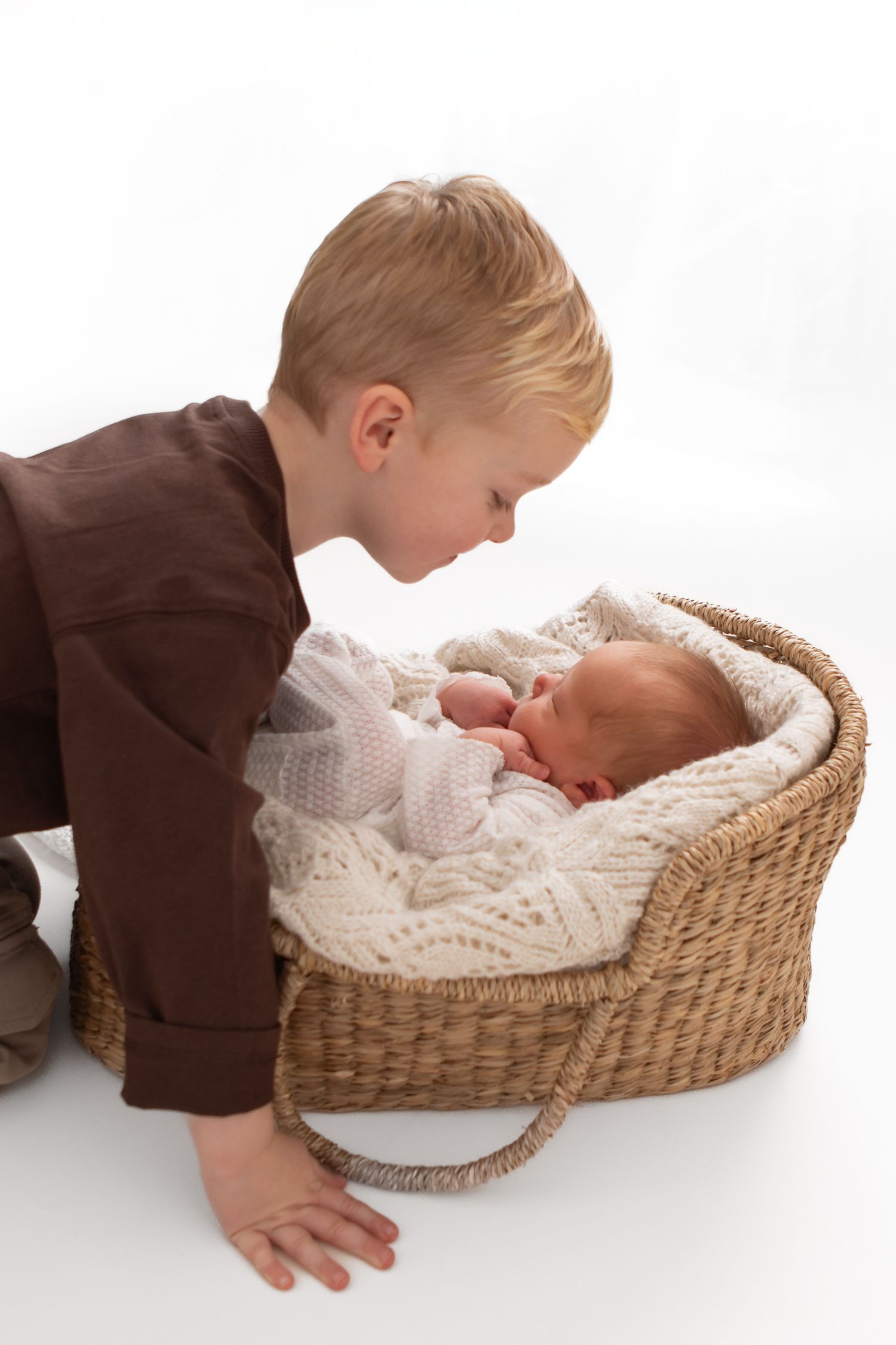Young child peering into woven basket containing sleeping newborn on white background.