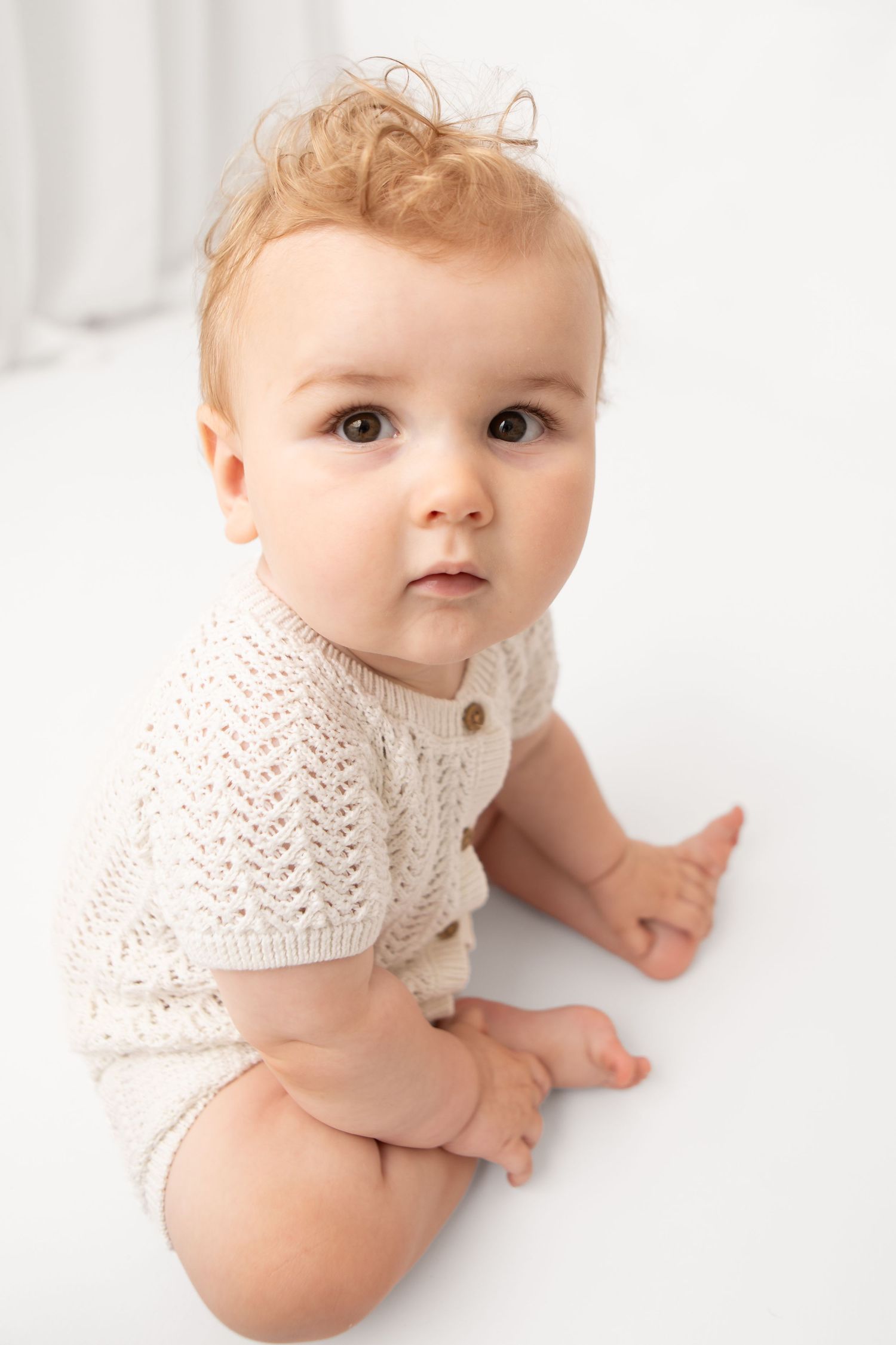 Adorable baby with curly red hair wearing a cream knit romper sitting on a white floor looking at camera.
