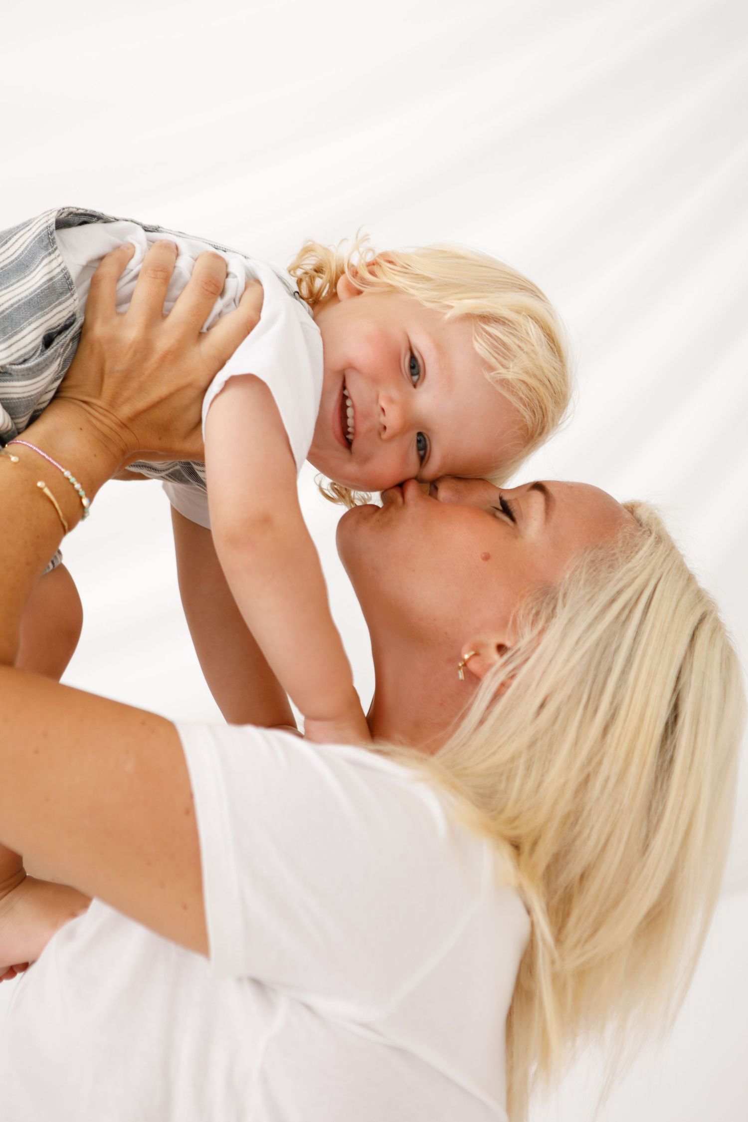 A joyful blonde mother lifting and kissing her smiling toddler against a white background.