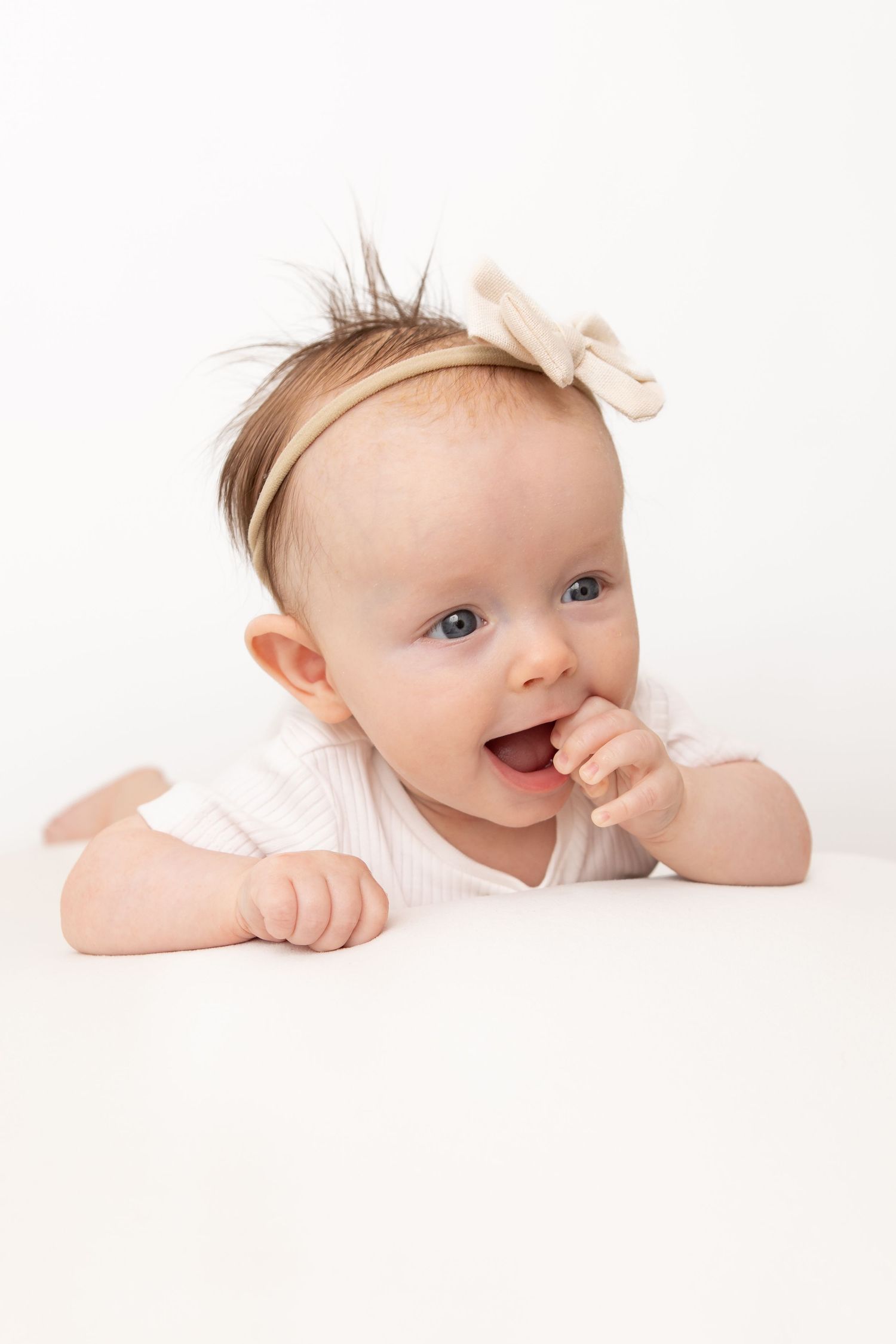 Adorable baby girl with a bow headband lying on her tummy, smiling with finger in mouth on white background.