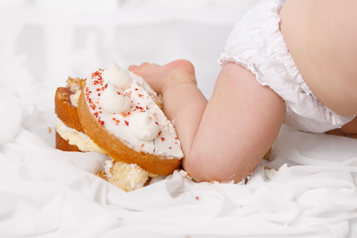 Baby feet holding a cream-filled pastry with white frosting and red sprinkles on a white background.