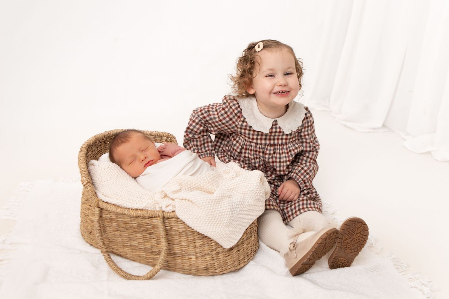 Toddler in brown dress sits near woven basket containing sleeping baby.