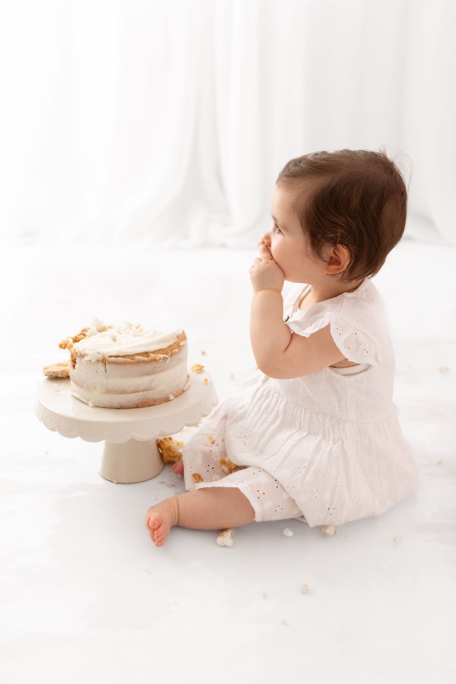 Baby girl in white dress eating cake at her first birthday smash cake photo session.