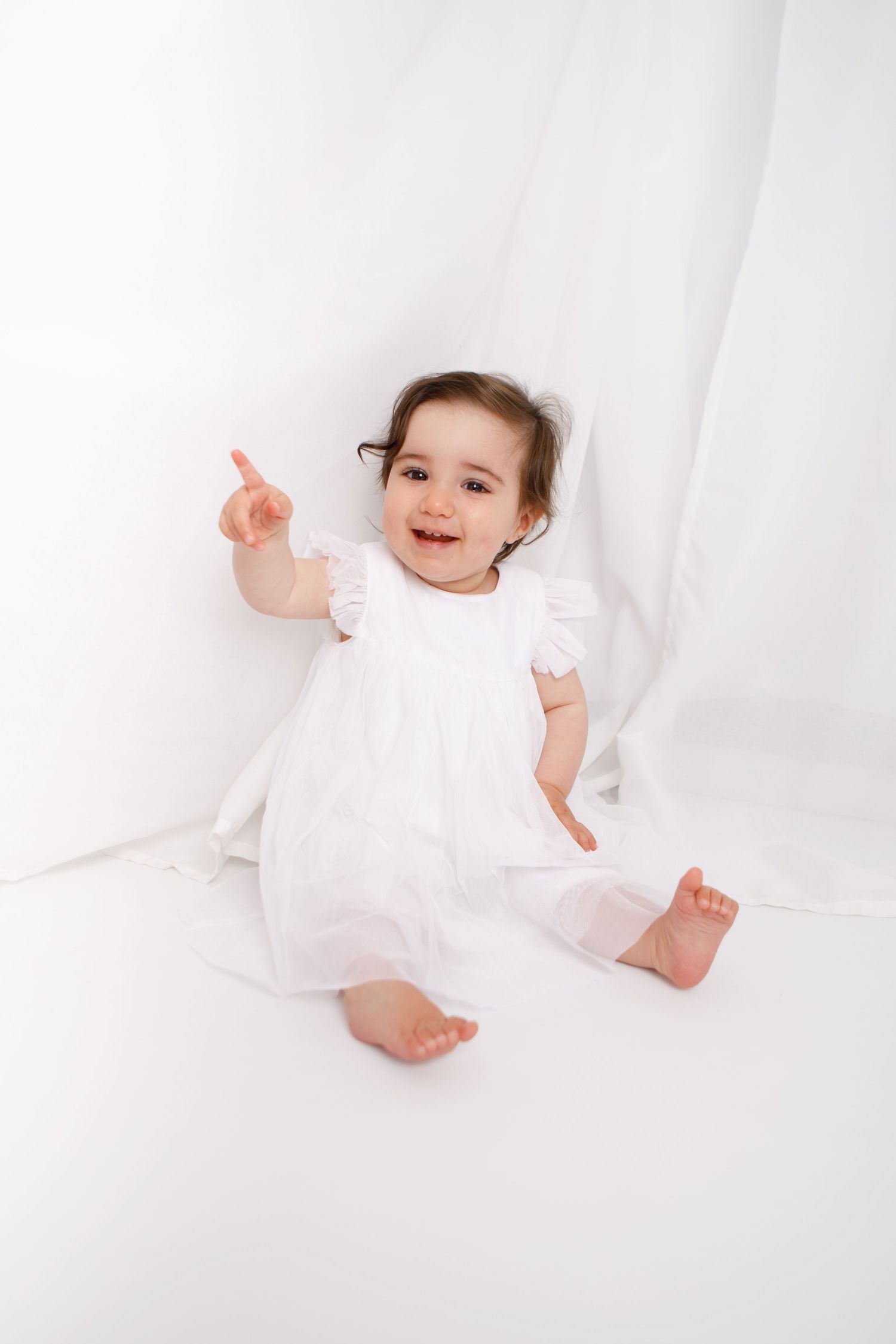 Baby in white dress lying on white backdrop with playful poses.
