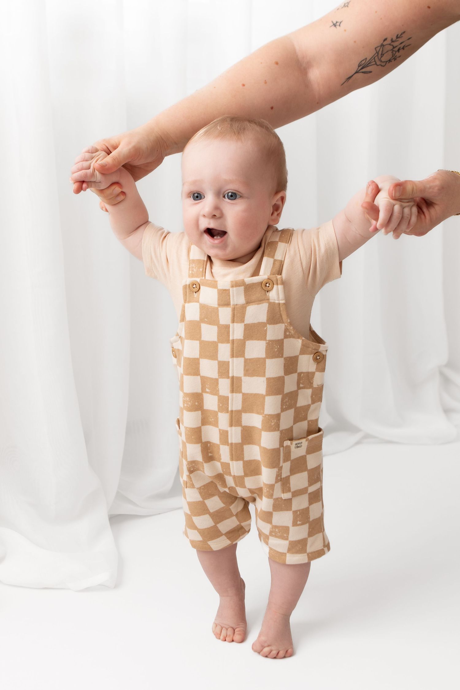 Baby in checkered tan overalls stands with adult hands supporting, smiling against white curtain backdrop.
