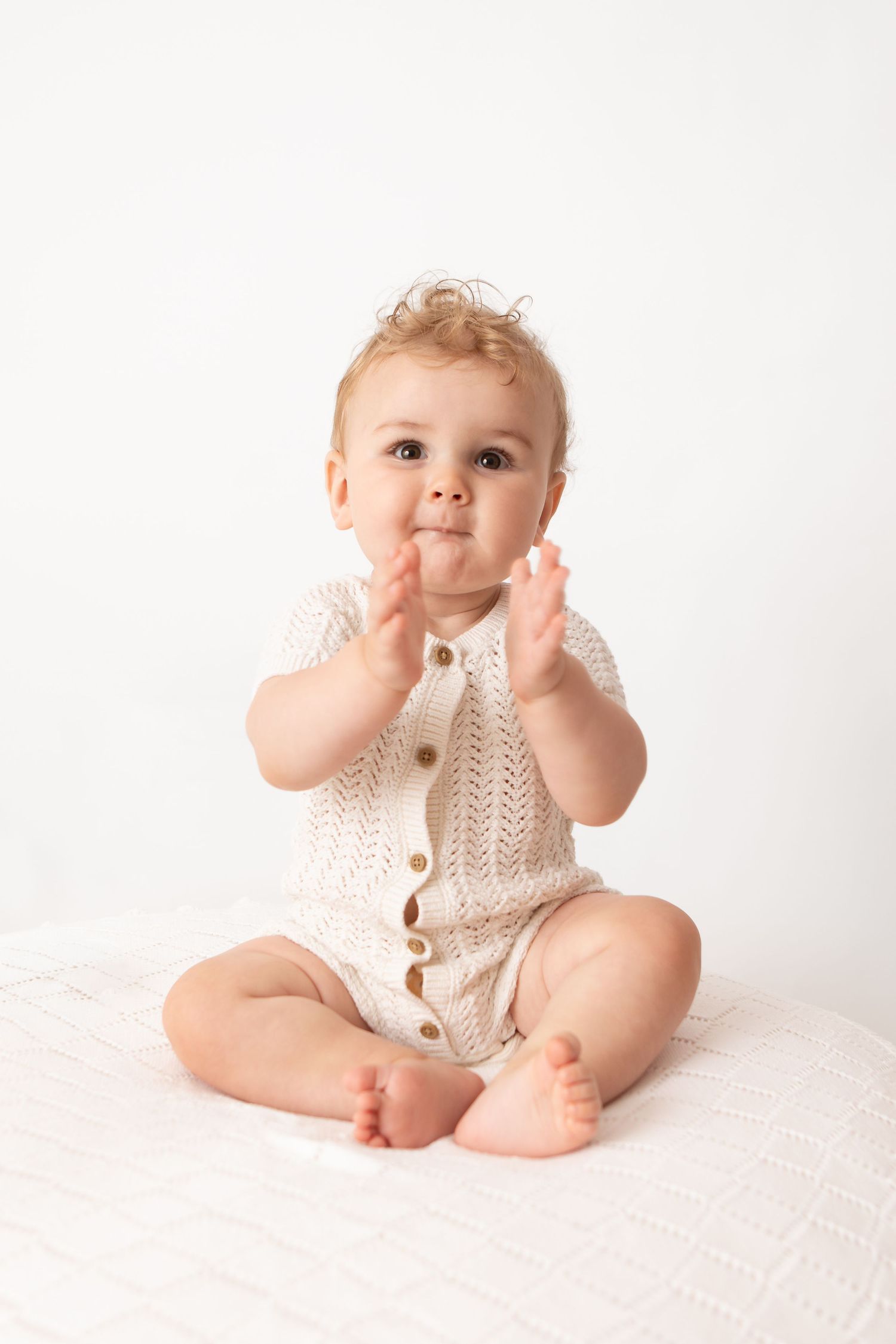Adorable baby in a cream knit romper sitting barefoot, clapping hands and looking at camera on white background.