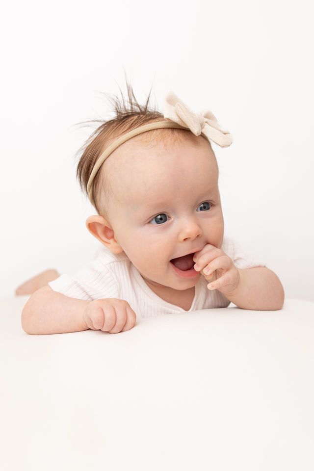 Adorable baby girl with a bow headband lying on her tummy, smiling with finger in mouth on white background.