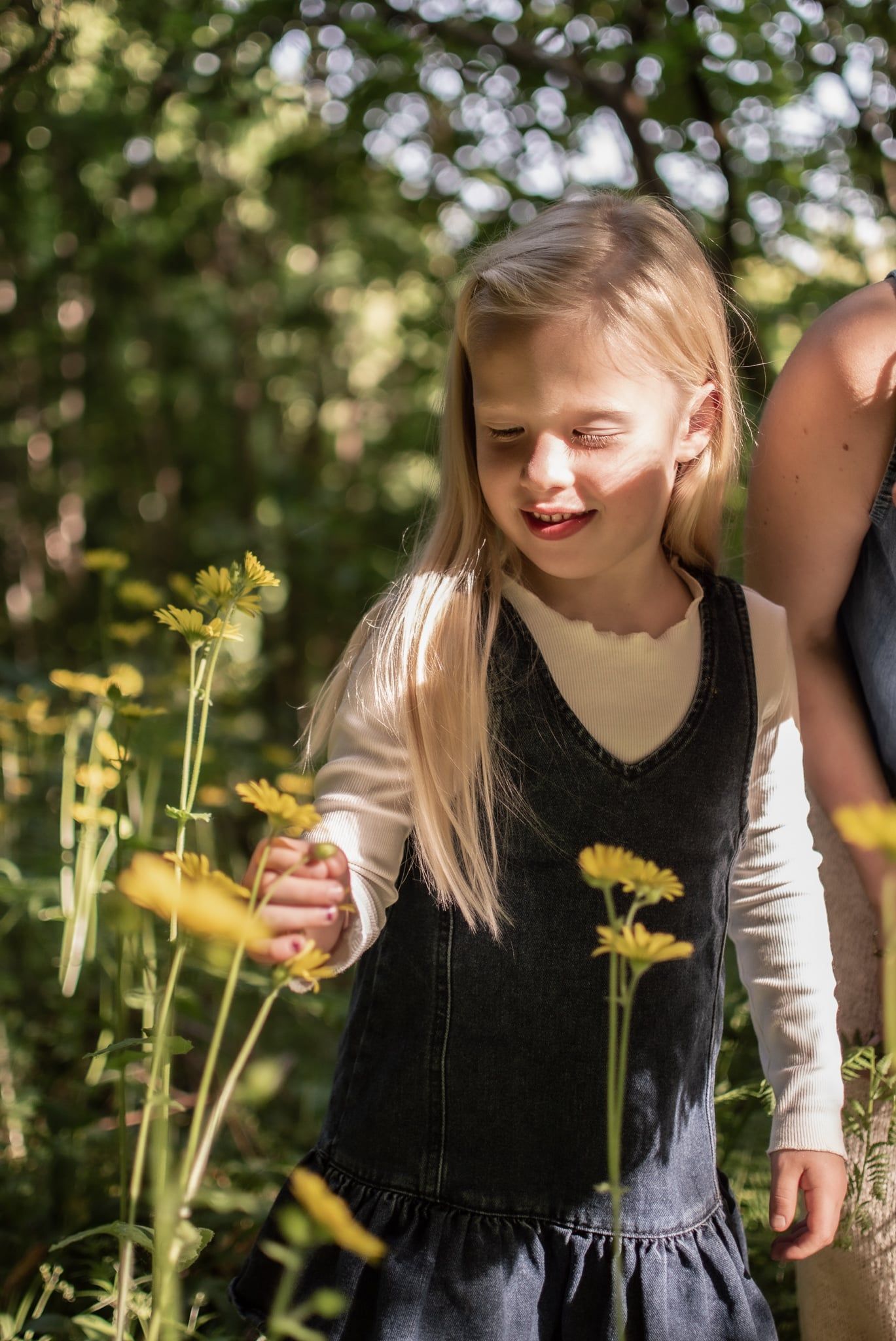 A smiling blonde girl reaches for yellow wildflowers in a sunny garden.