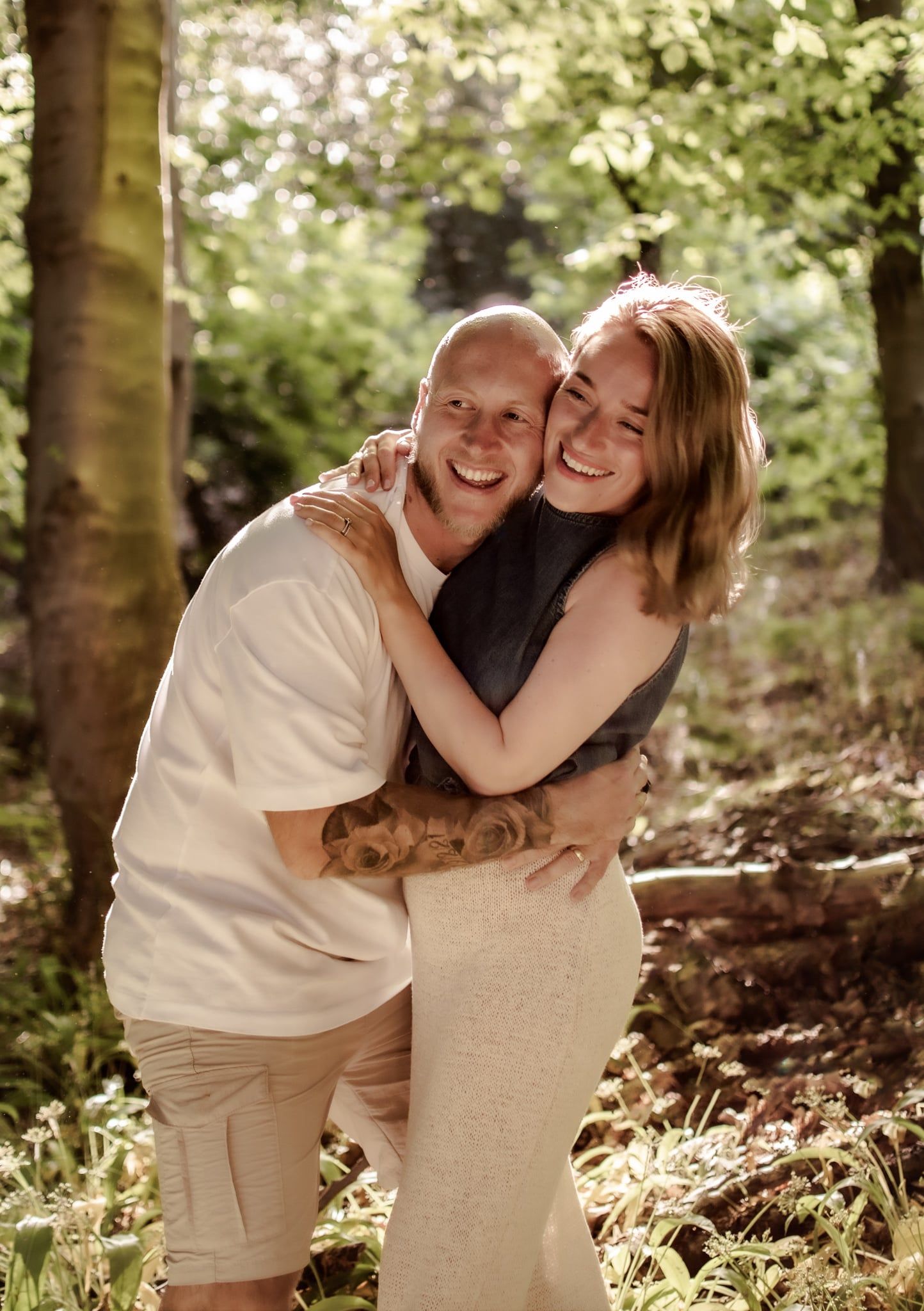 A smiling couple embraces warmly in a sunlit forest, the woman hugging the bald man from behind among green trees.
