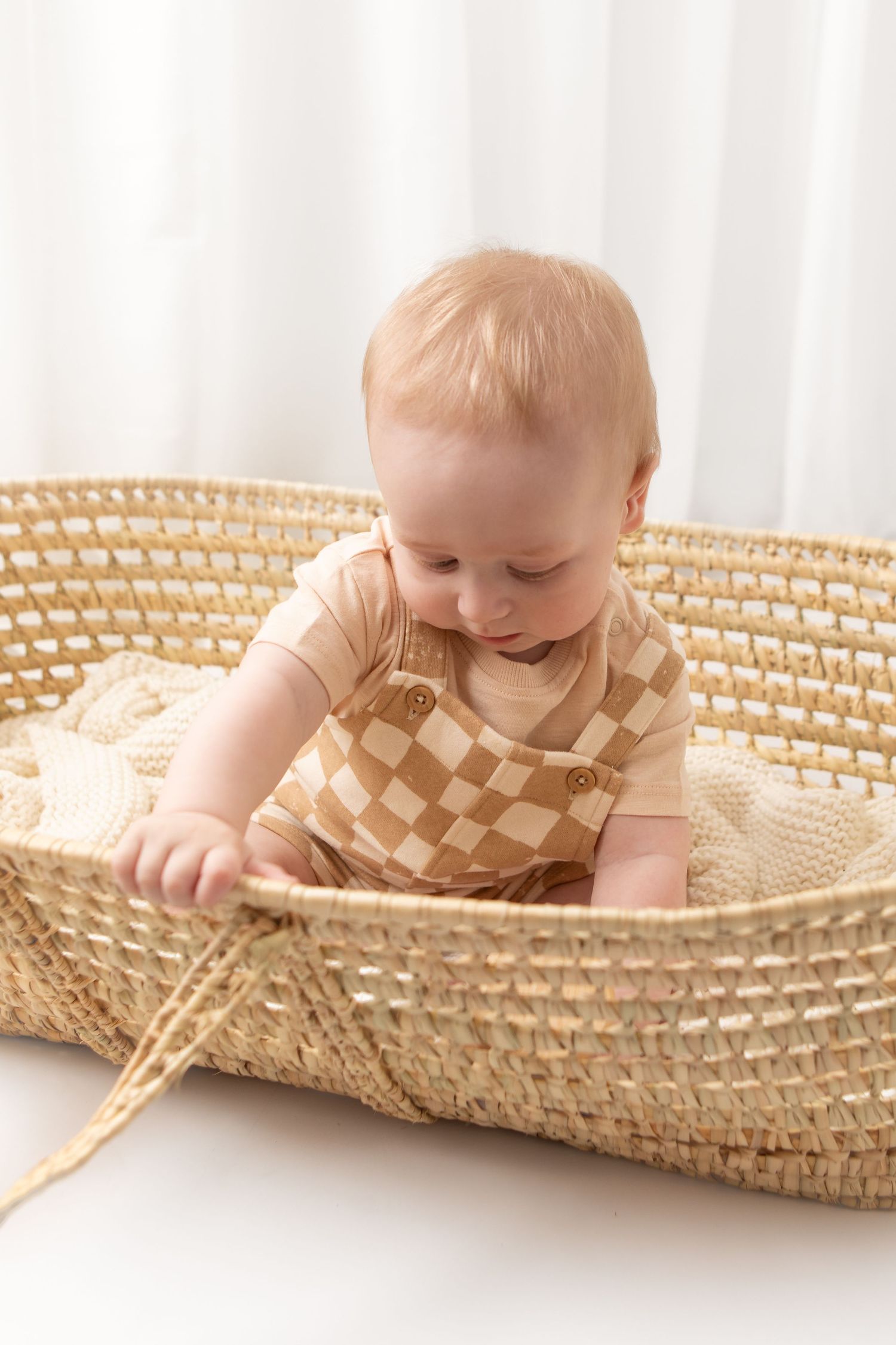 Baby in checkered romper sitting in a woven Moses basket, looking down at the basket edge.