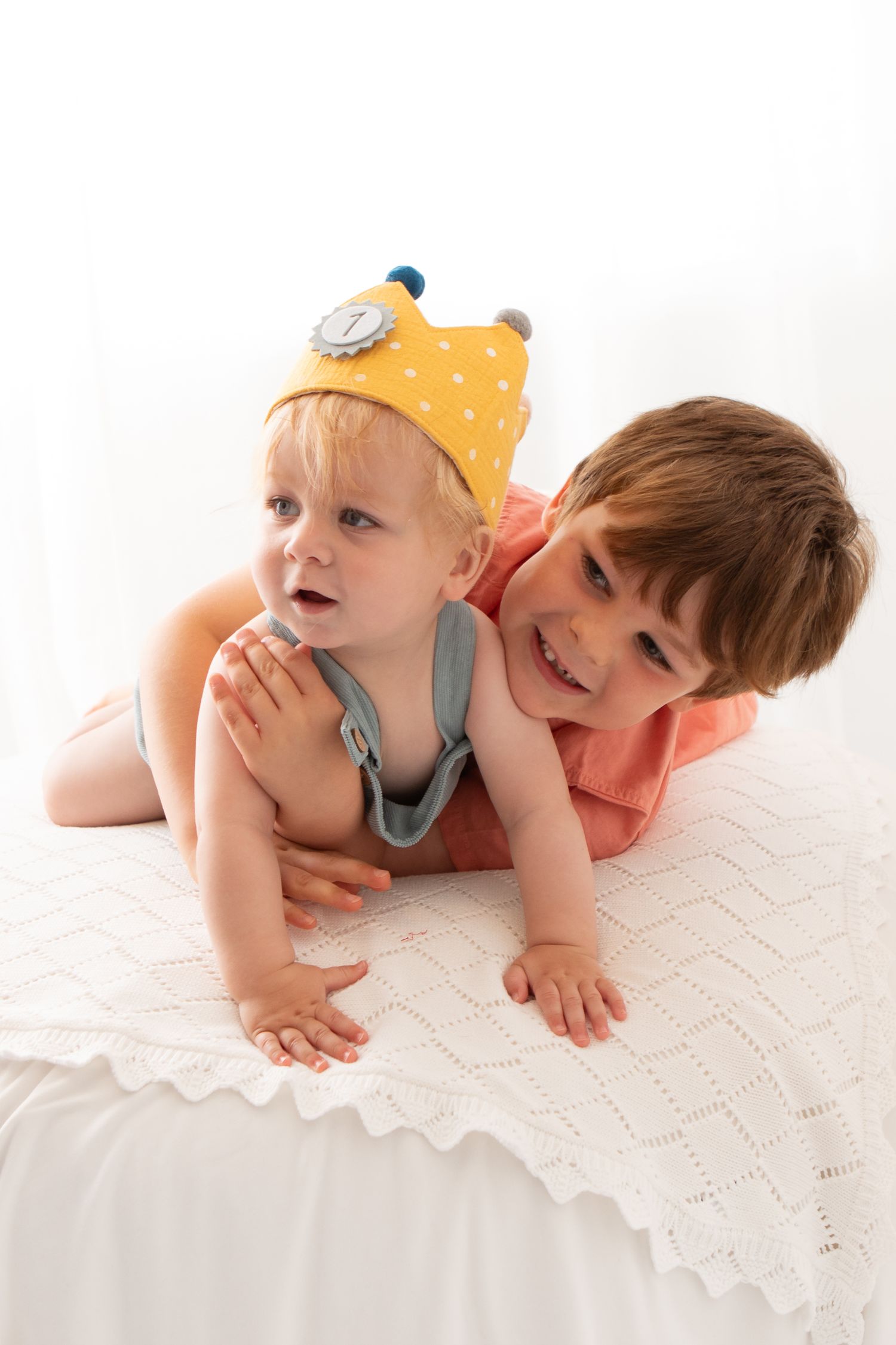 Two young siblings lying on a white blanket, older boy smiling behind baby wearing a yellow crown hat.