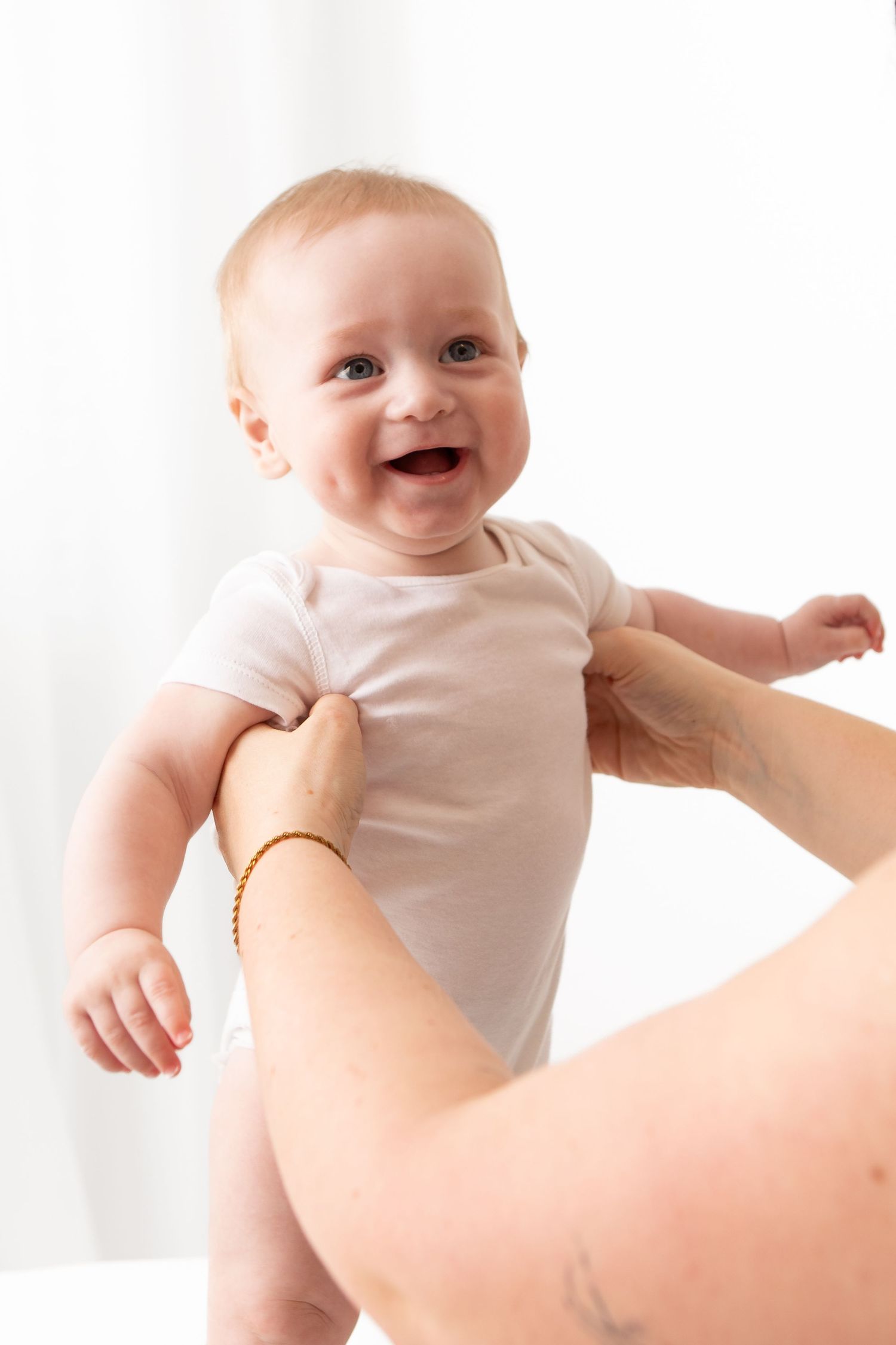 Smiling baby in white onesie being held up by adult hands with gold bracelet against white background.