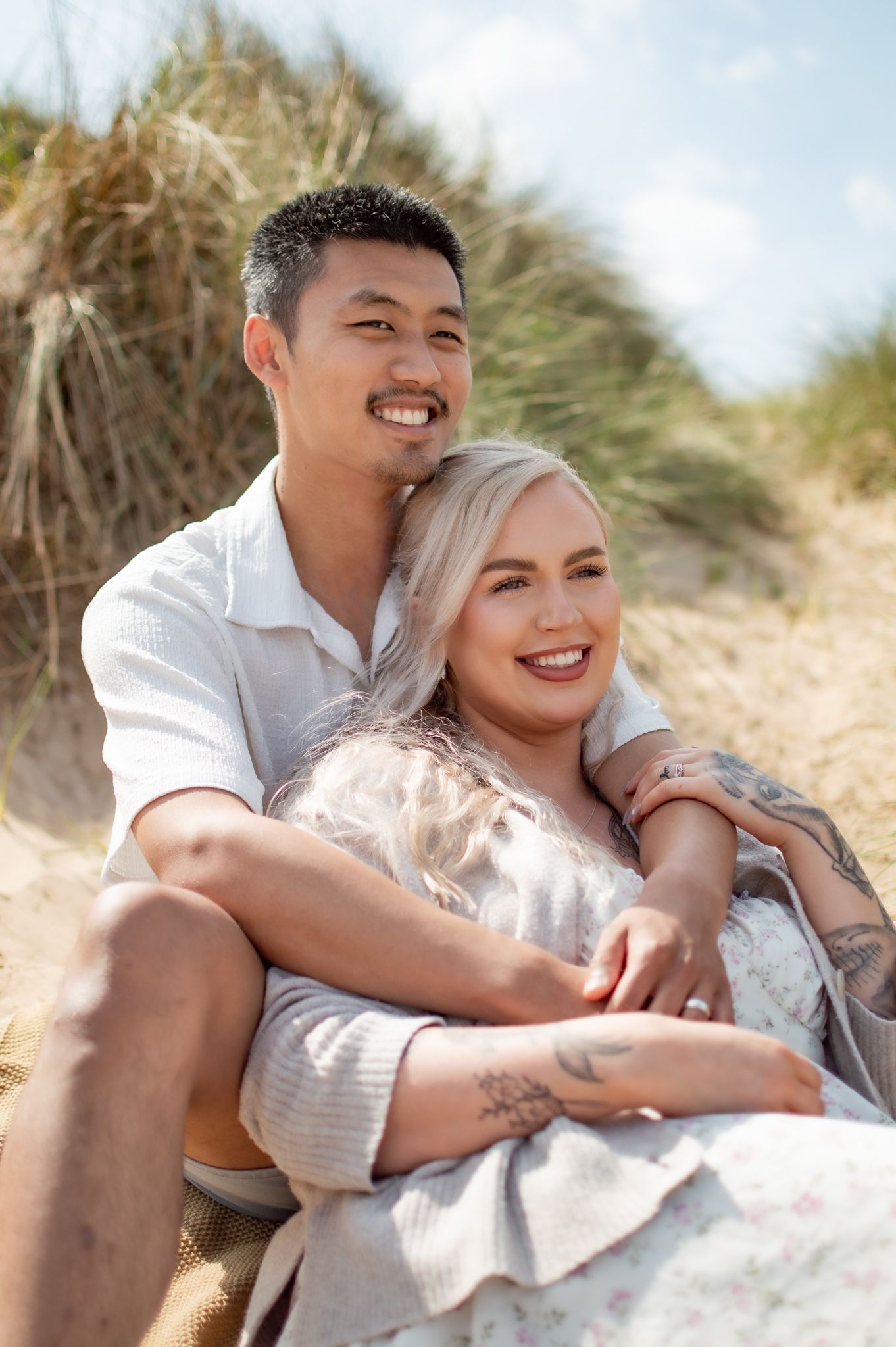 Couple smiling and embracing on sandy beach dunes, woman with blonde hair leaning against man in white shirt on sunny day.