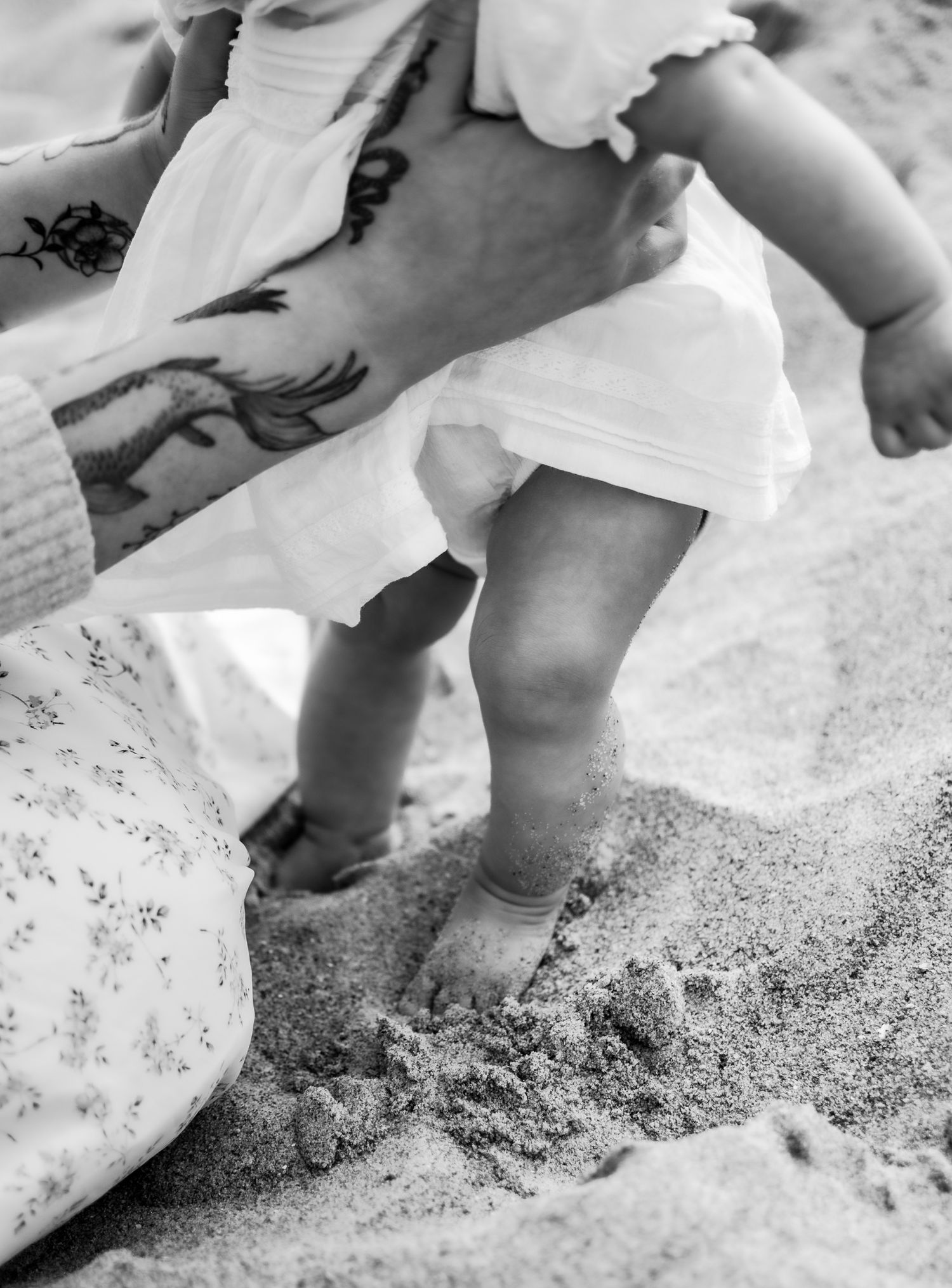 Close-up of baby feet touching sand at the beach, held by tattooed adult hands in black and white.