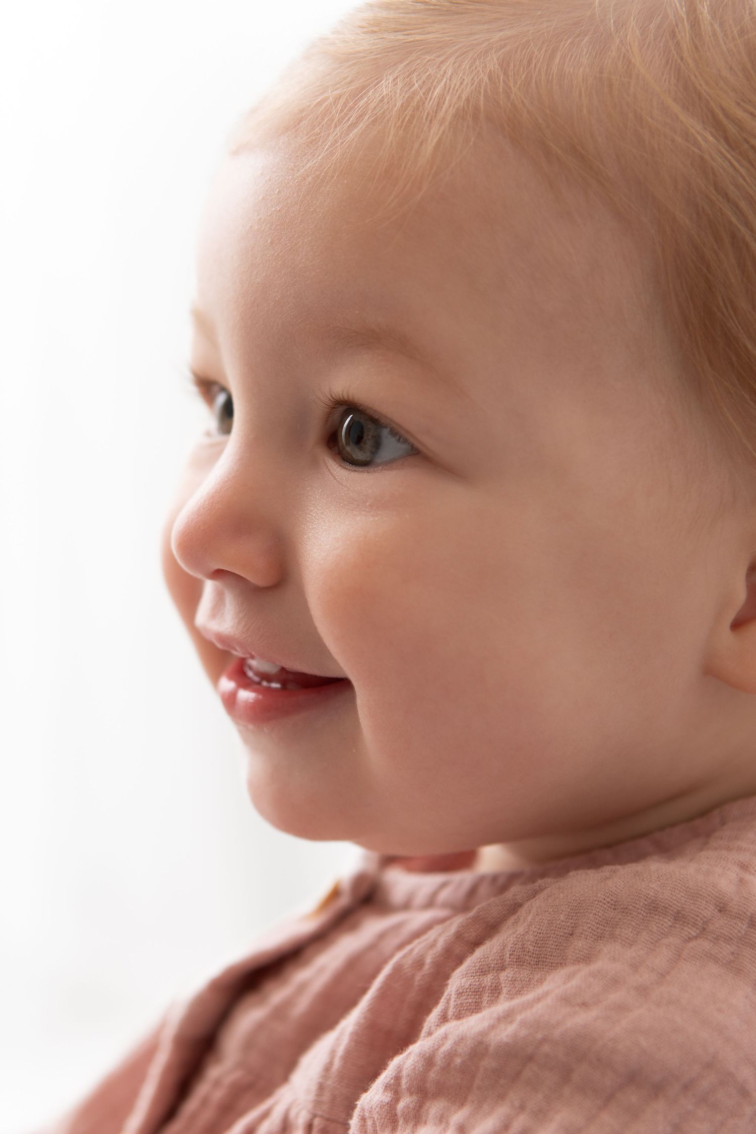 Smiling baby with rosy cheeks wearing a pink outfit, captured in a close-up portrait against a white background.