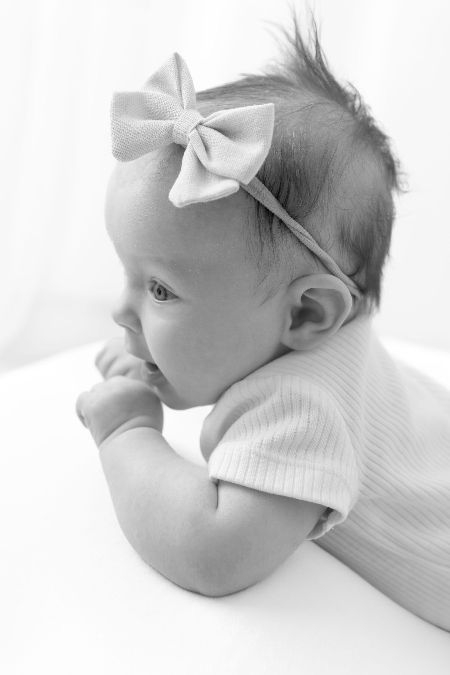 Black and white photo of a cute baby girl wearing a bow headband, resting on her tummy and looking sideways.