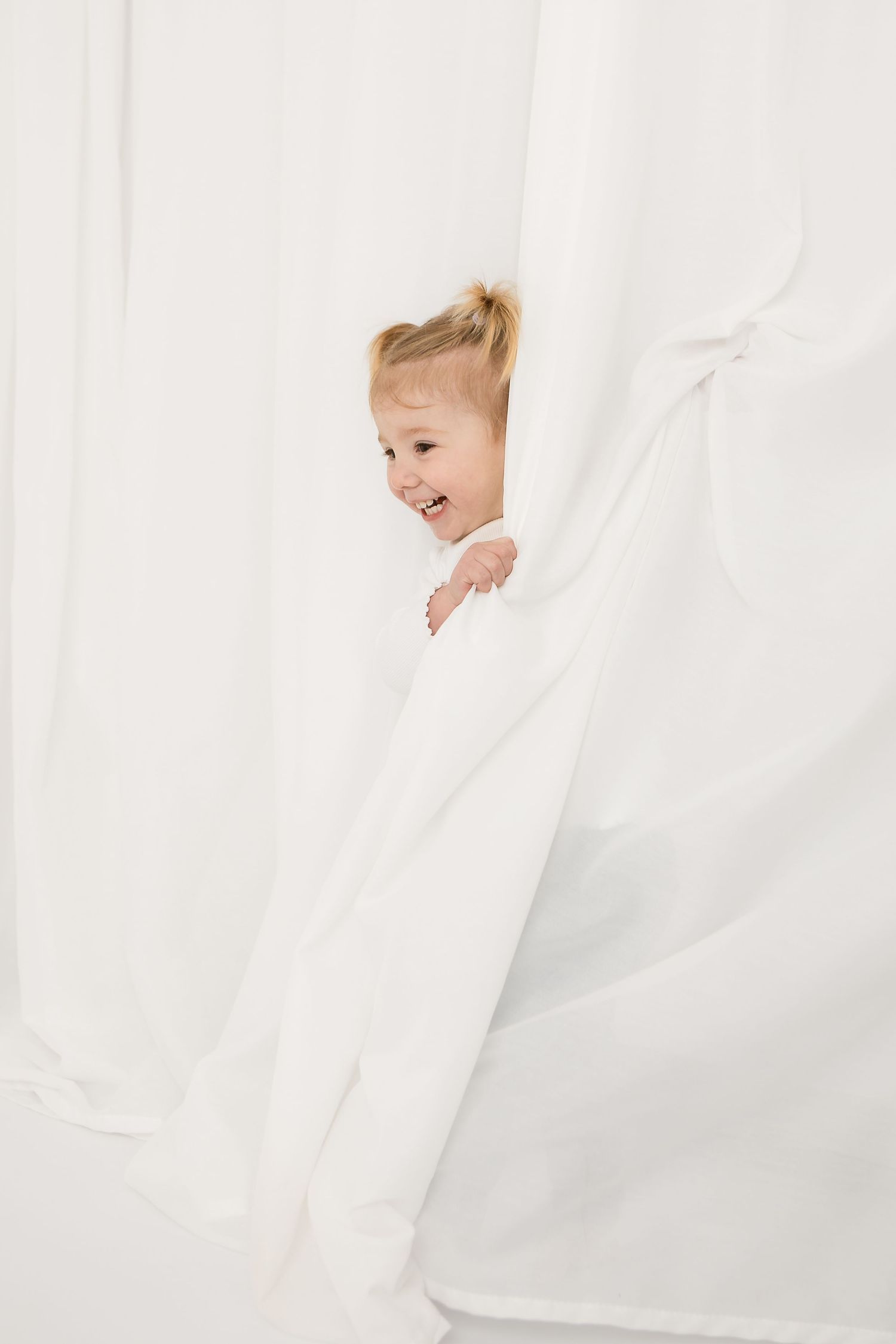 Joyful toddler girl peeking through white curtains, laughing and playing peek-a-boo in a bright white room.