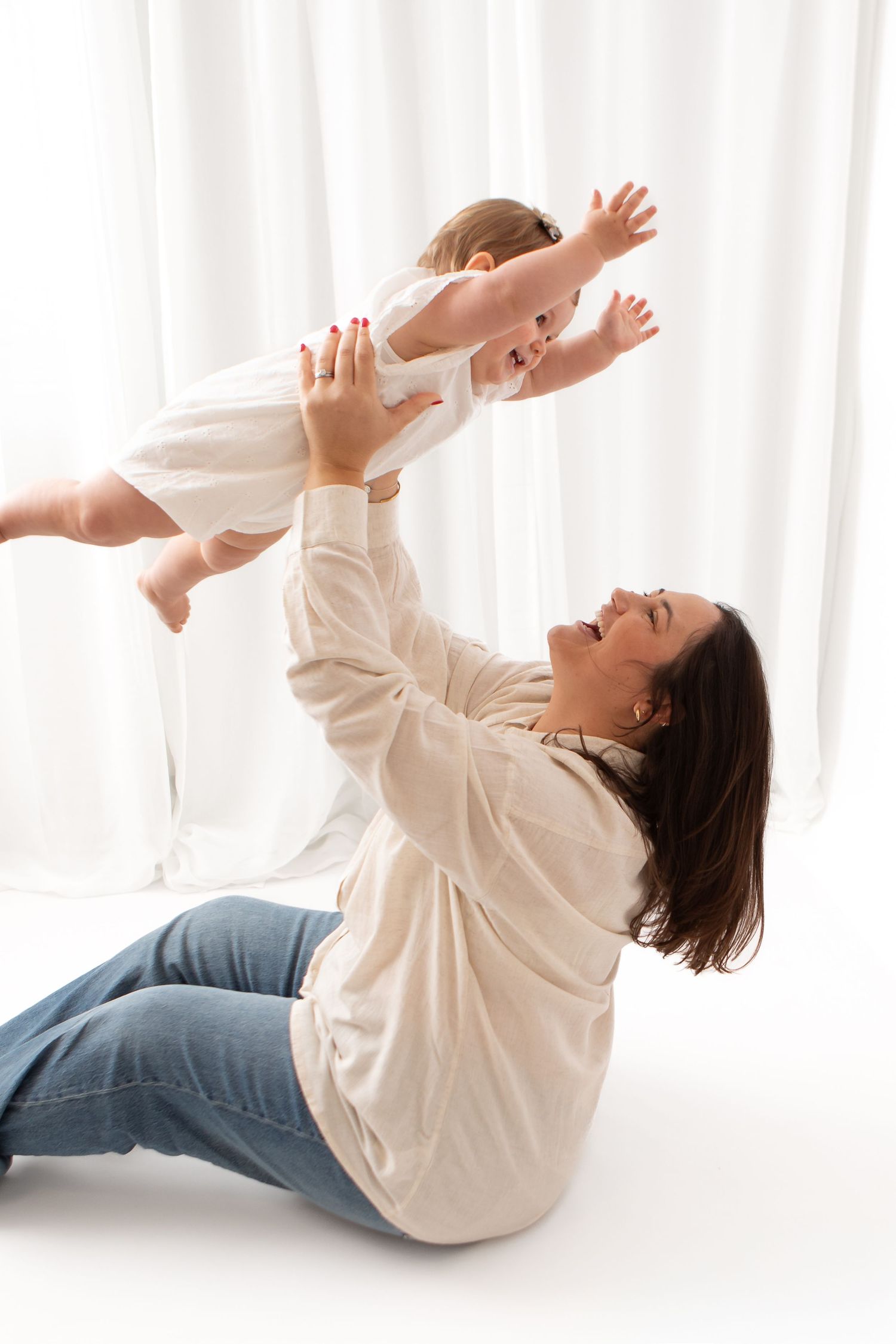 A mother sits on the floor lifting her smiling baby girl overhead, both enjoying playful bonding time together.