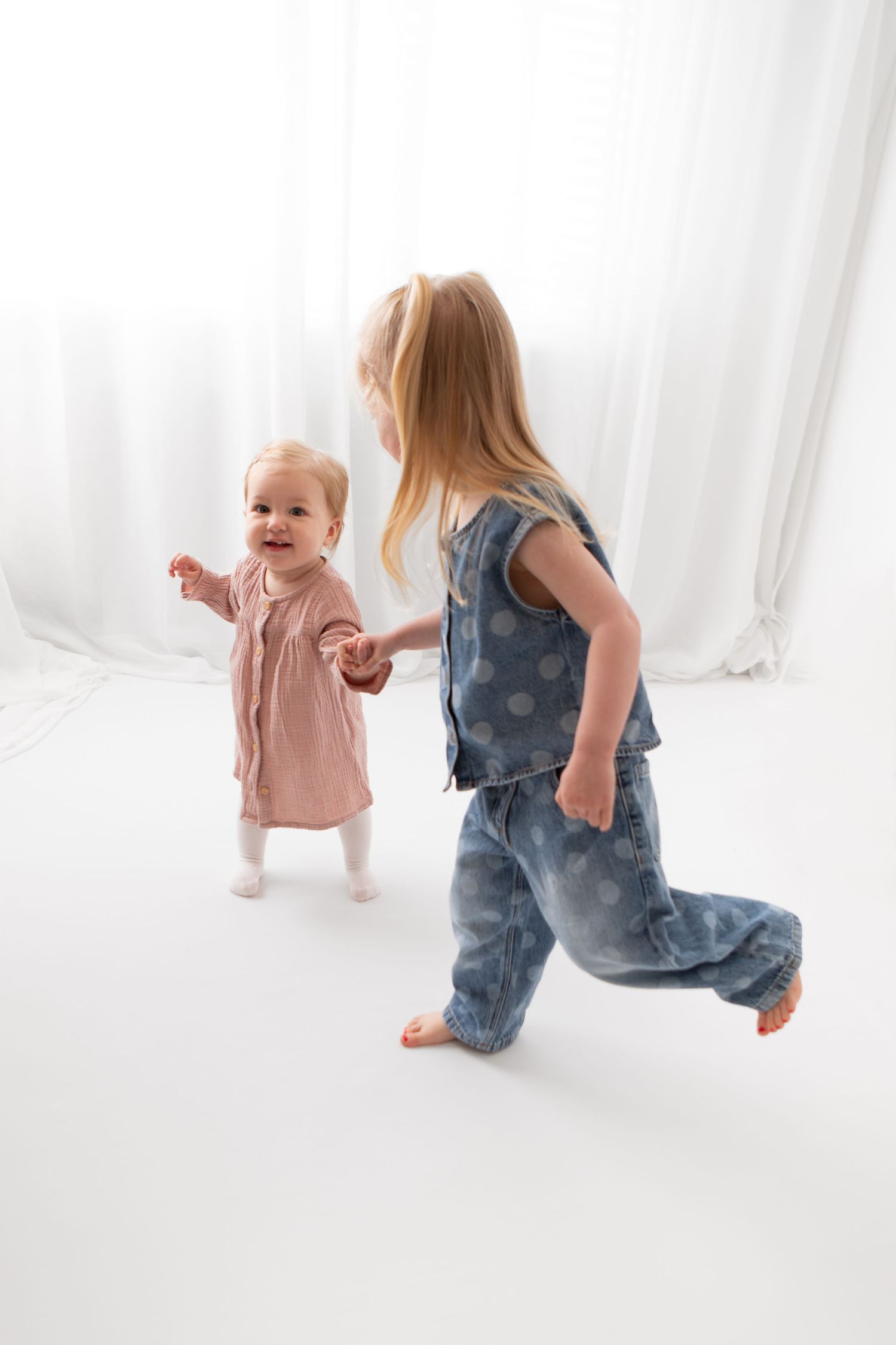 Two young girls holding hands and playing together, wearing matching blue polka dot and pink outfits in a bright white studio.