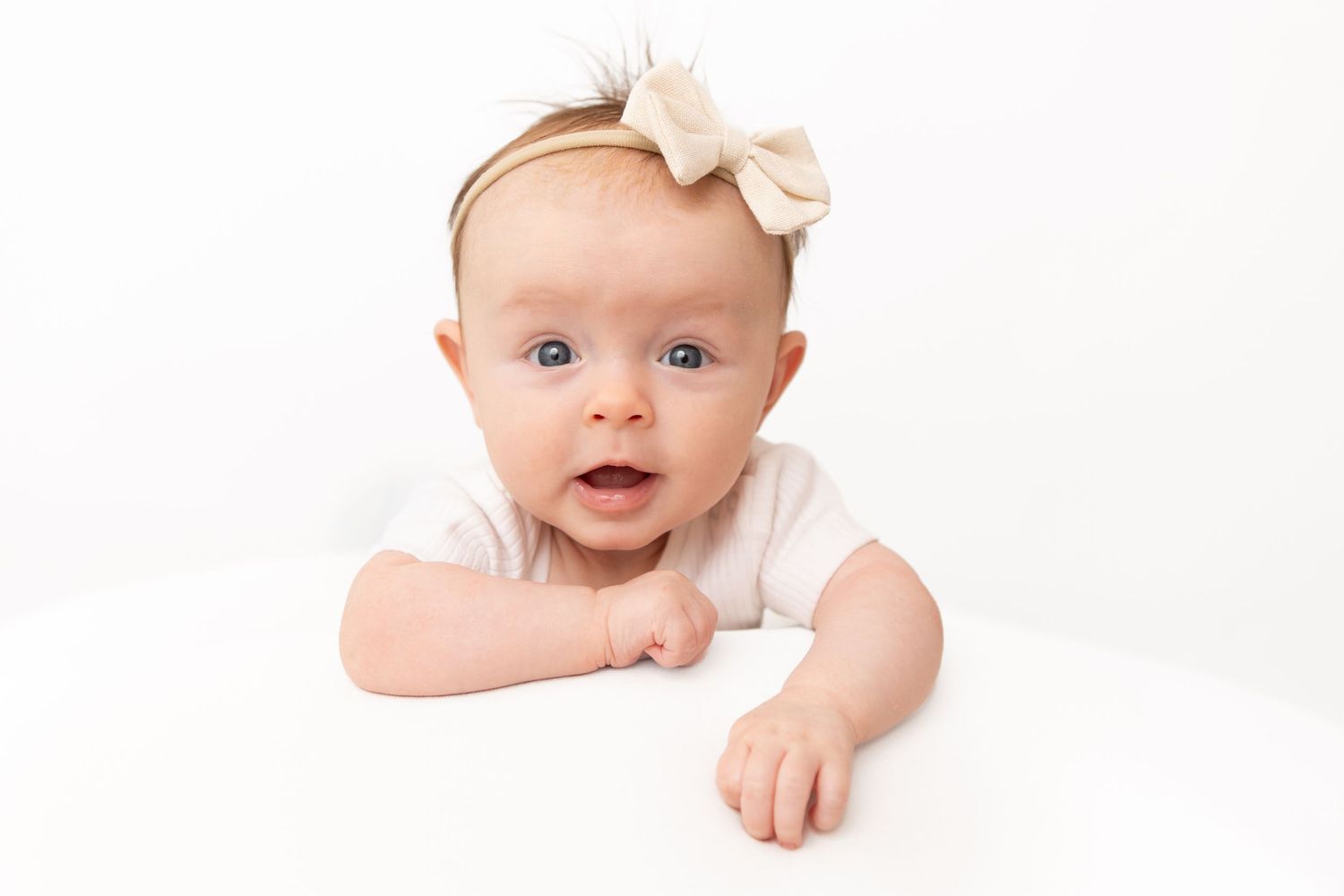 Adorable baby girl with blue eyes wearing a white onesie and bow headband doing tummy time on white background.