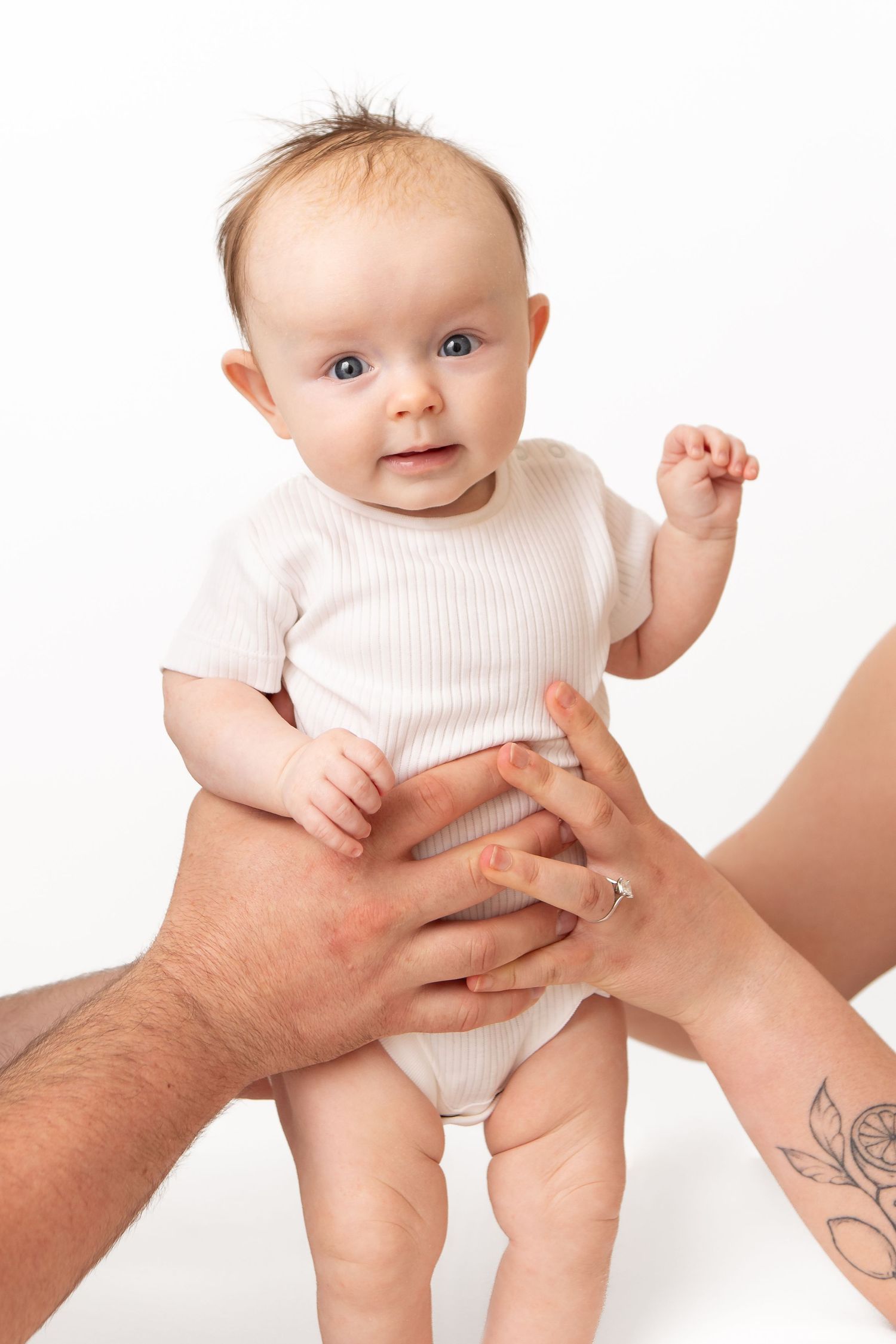 Adorable baby in white onesie held by parents hands, one with floral tattoo, against white background.