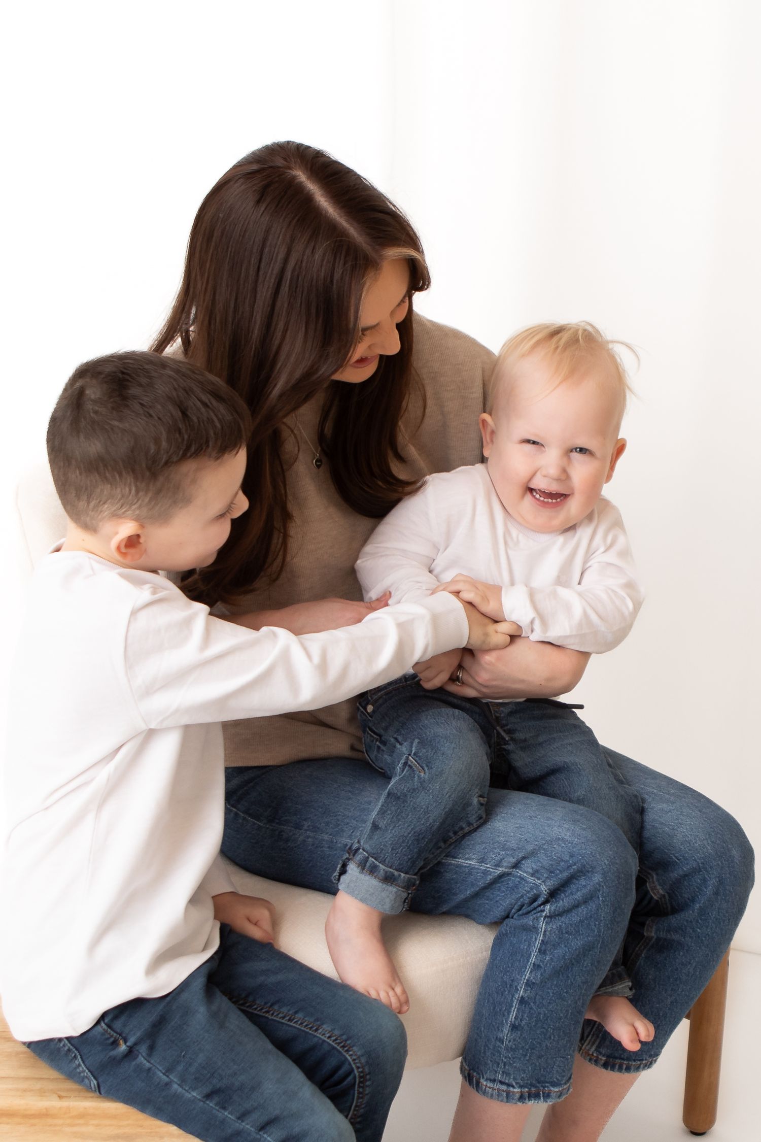 A smiling mother sits with her two young children, a toddler laughing joyfully while an older boy plays with them.