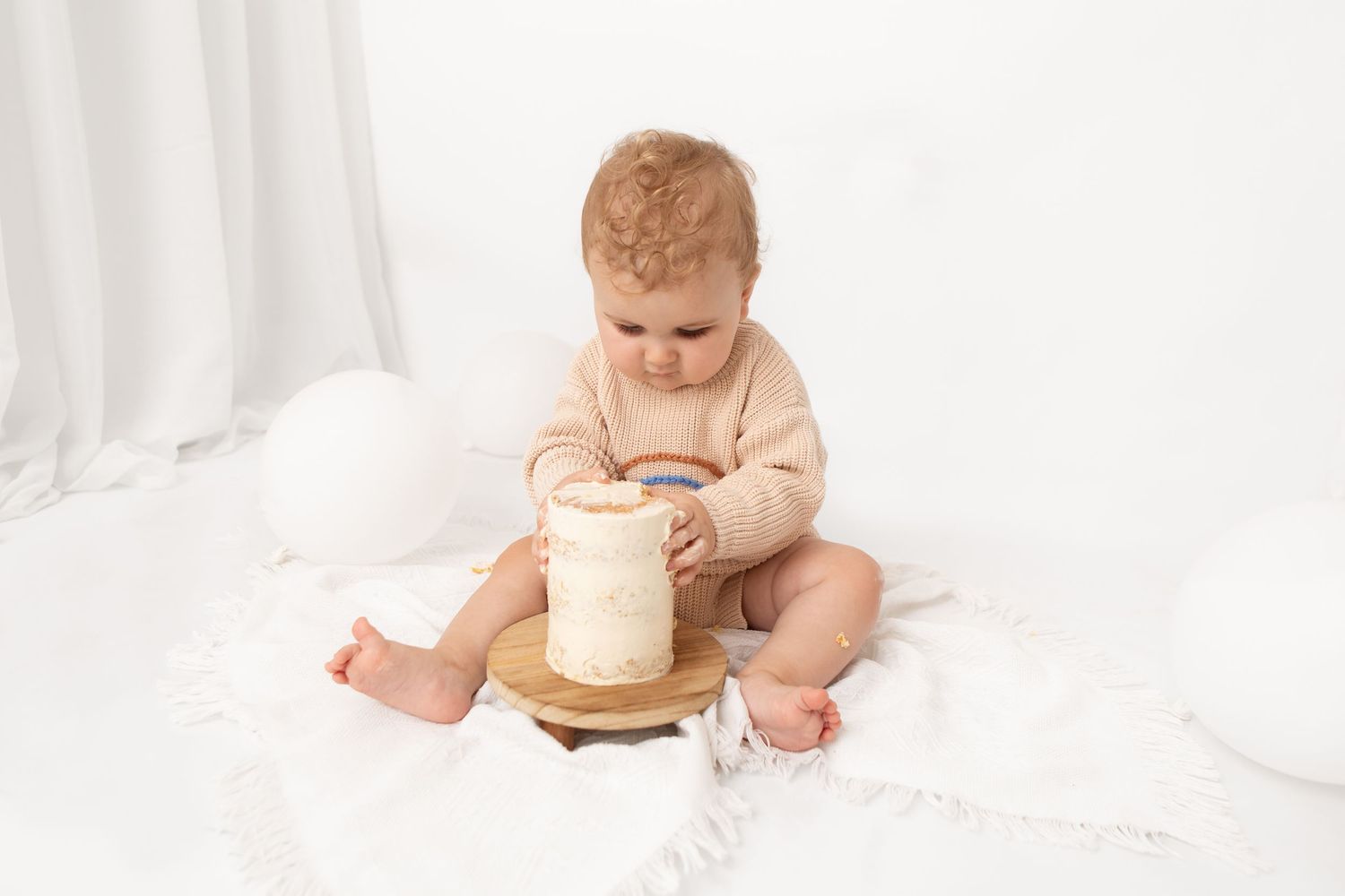 A baby in a beige romper sits on a white blanket exploring a small white cake on a wooden stand.