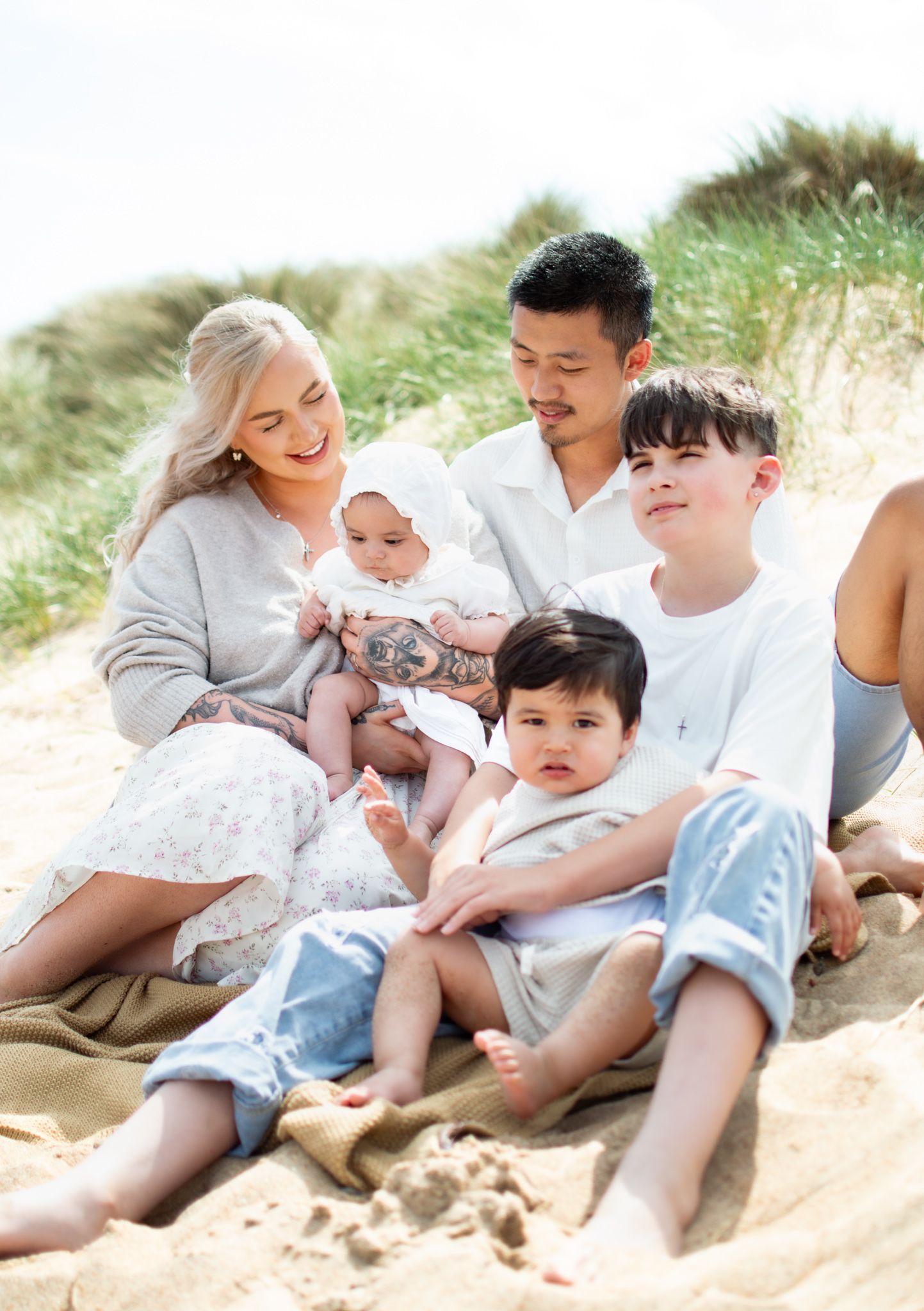 Family of five sitting on sandy beach dunes, parents with three young children enjoying a sunny day outdoors.