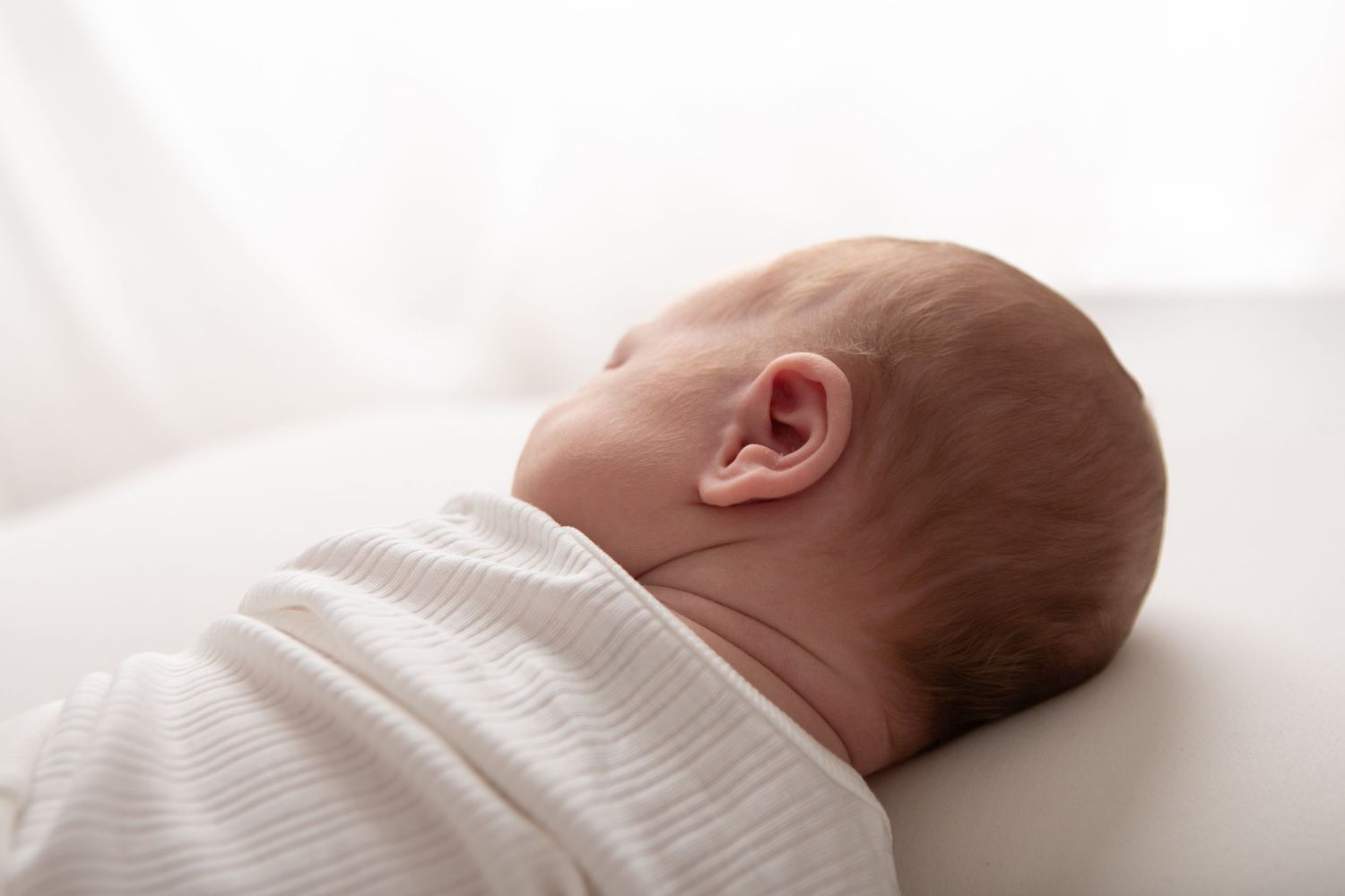 Newborn baby swaddled in white blanket sleeping peacefully on soft white surface, side profile view.