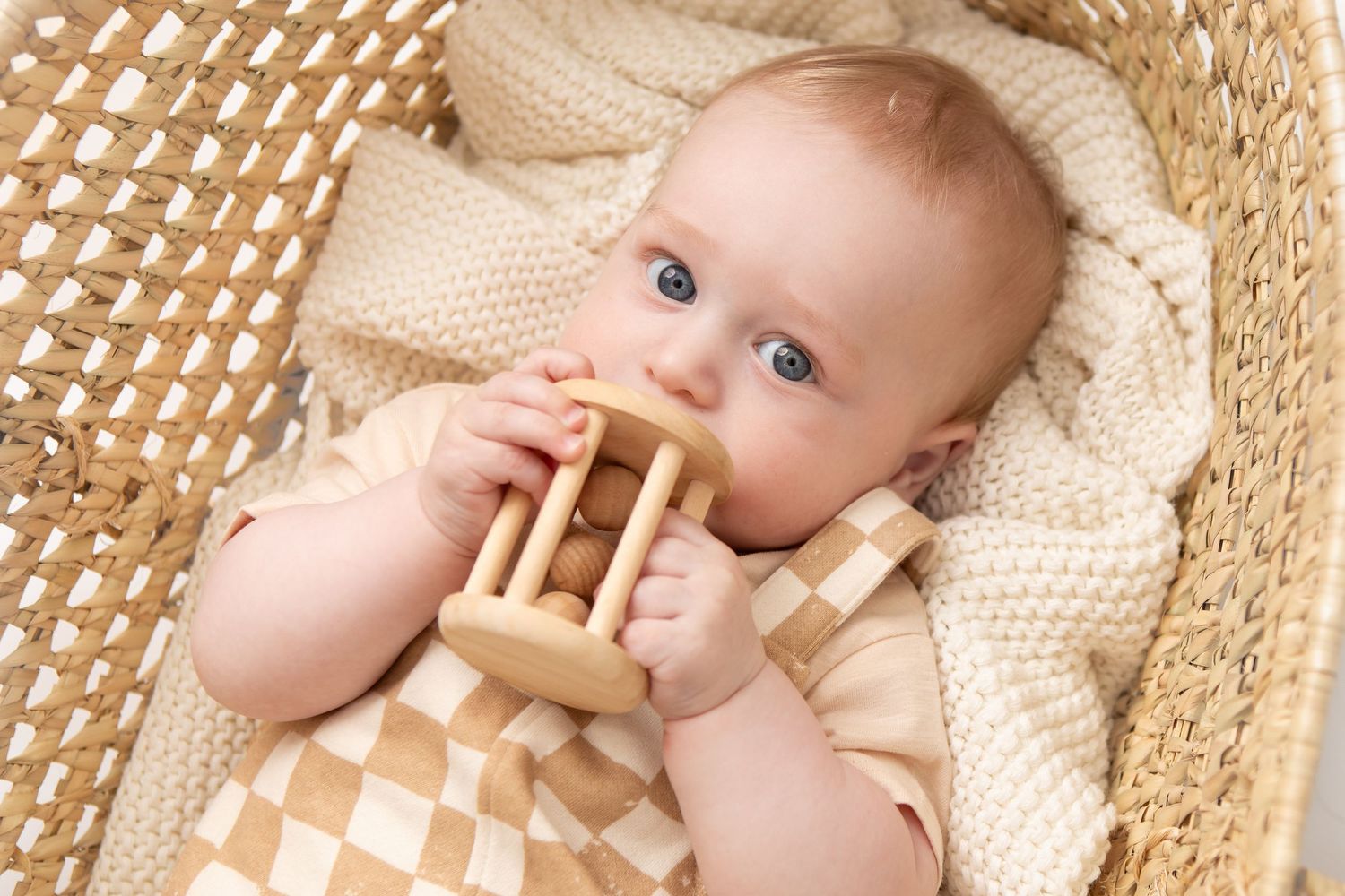 Baby with blue eyes chewing on a wooden toy rattle while lying in a wicker basket with a knit blanket.