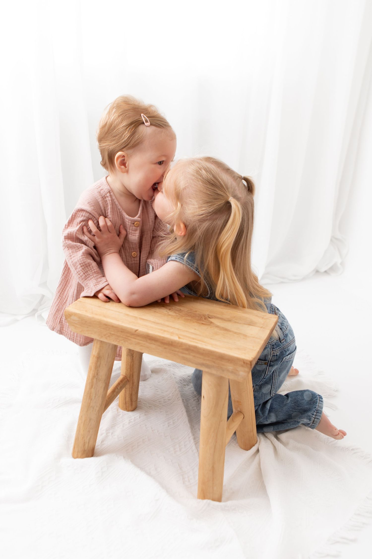 Two young girls sharing a sweet moment together beside a wooden step stool on a white background.