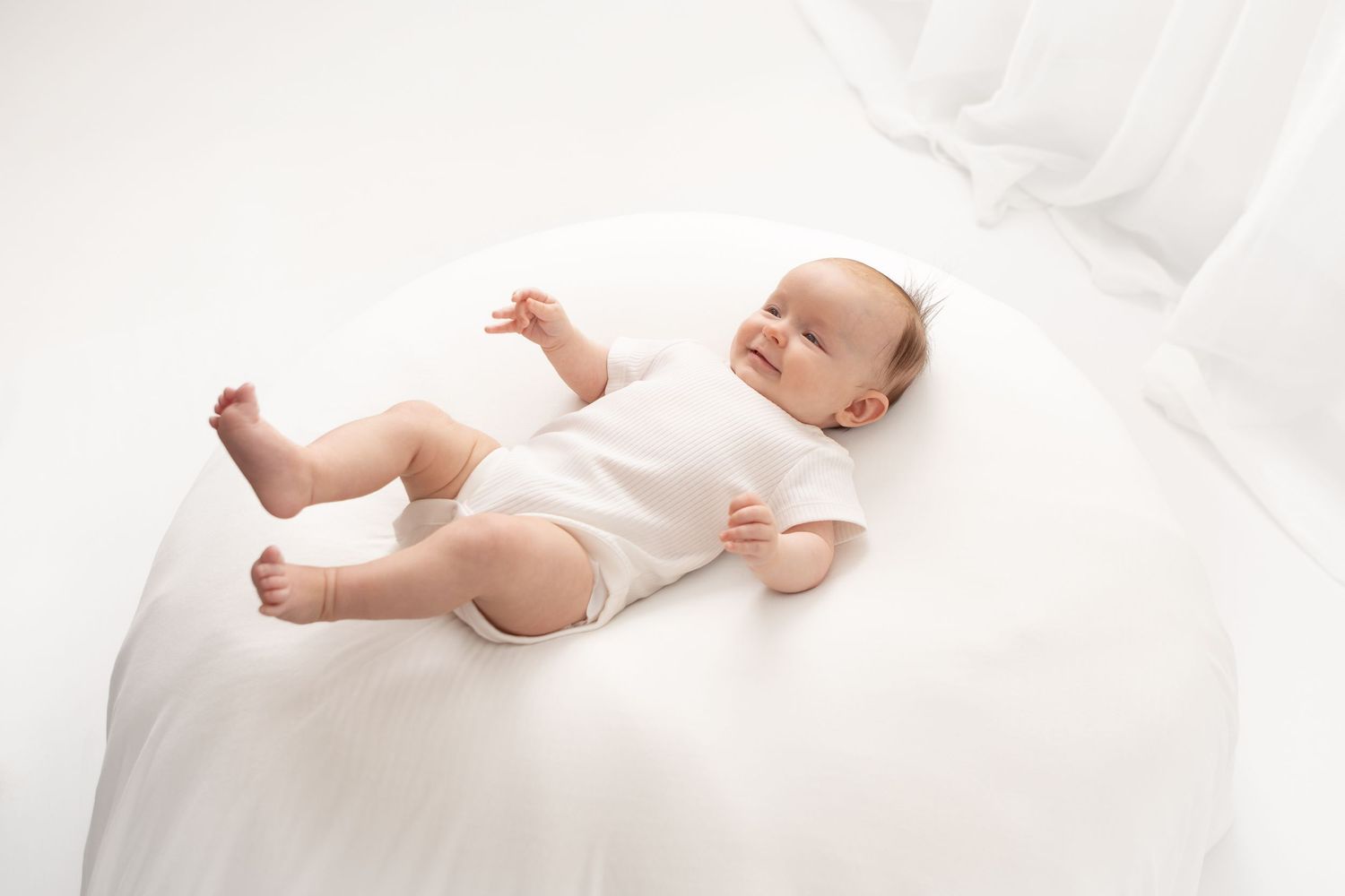 Smiling baby in white onesie lying on a white bed, looking content and relaxed in a bright, airy setting.