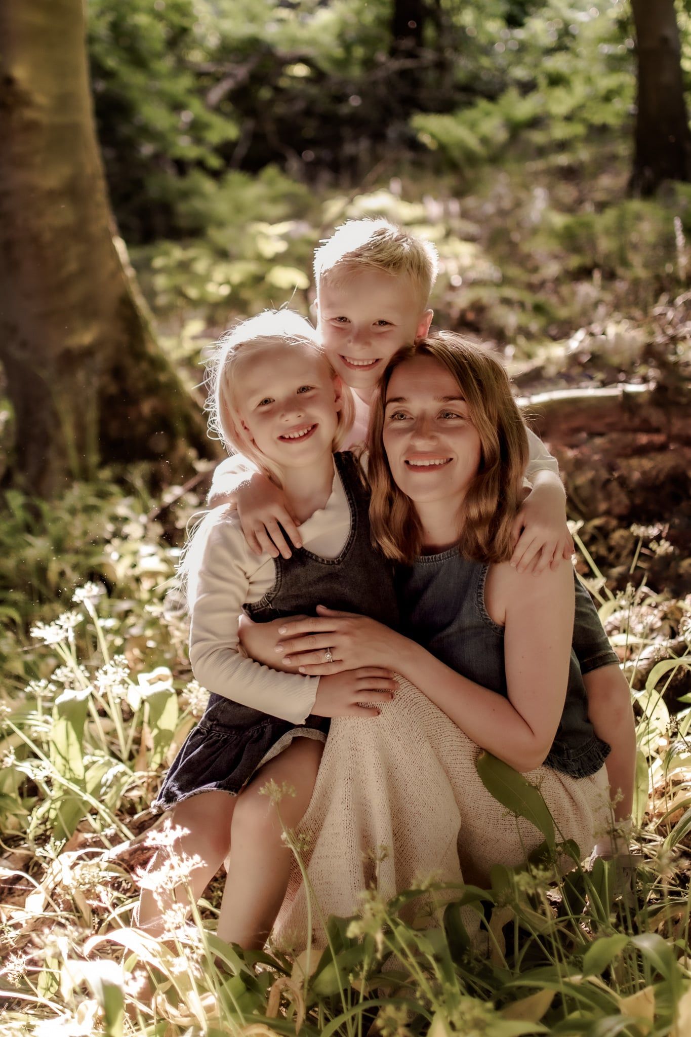 Three smiling children hugging in a sunlit forest, surrounded by green plants and wildflowers.