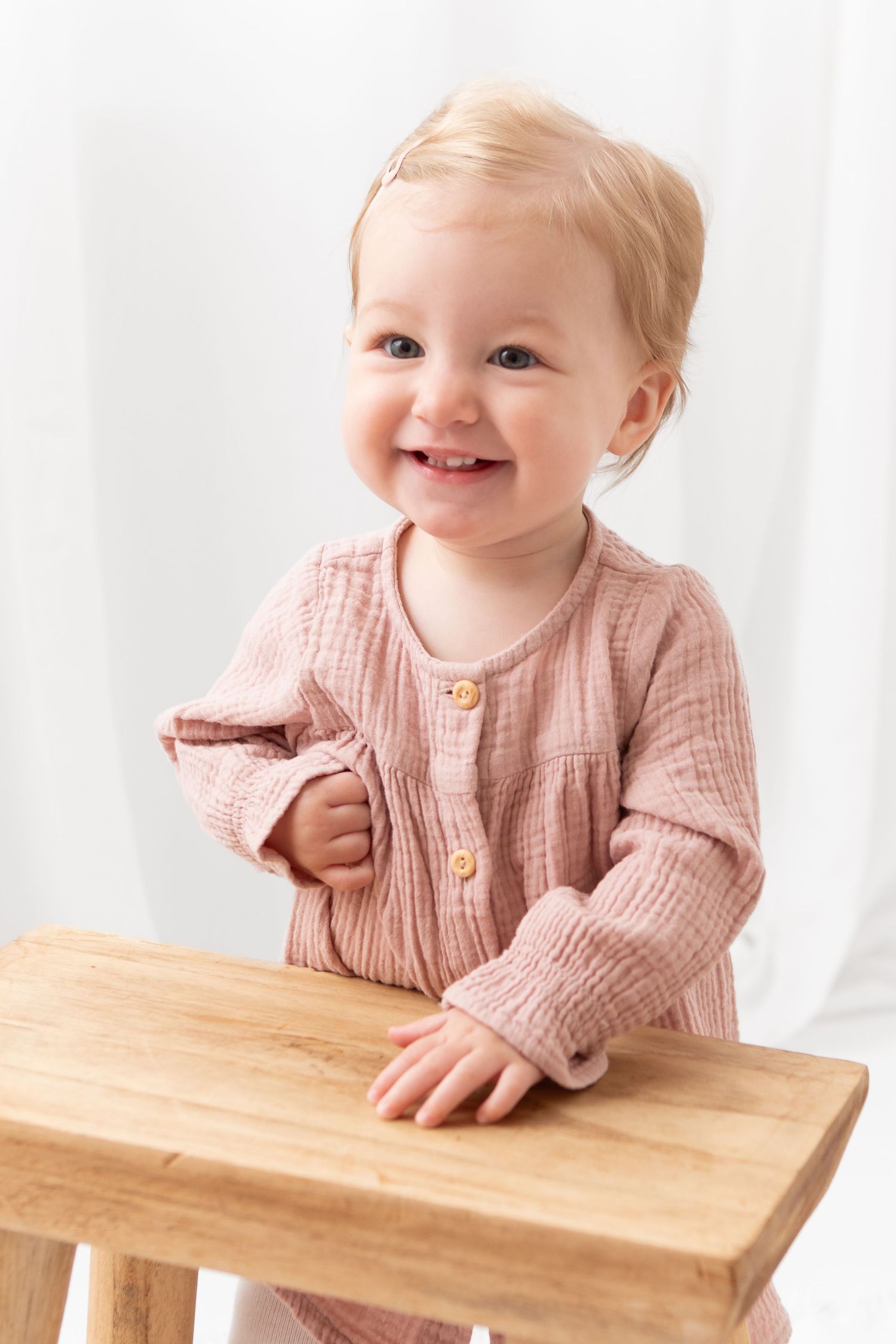 Smiling baby girl in pink muslin outfit standing at a wooden stool against a white background.