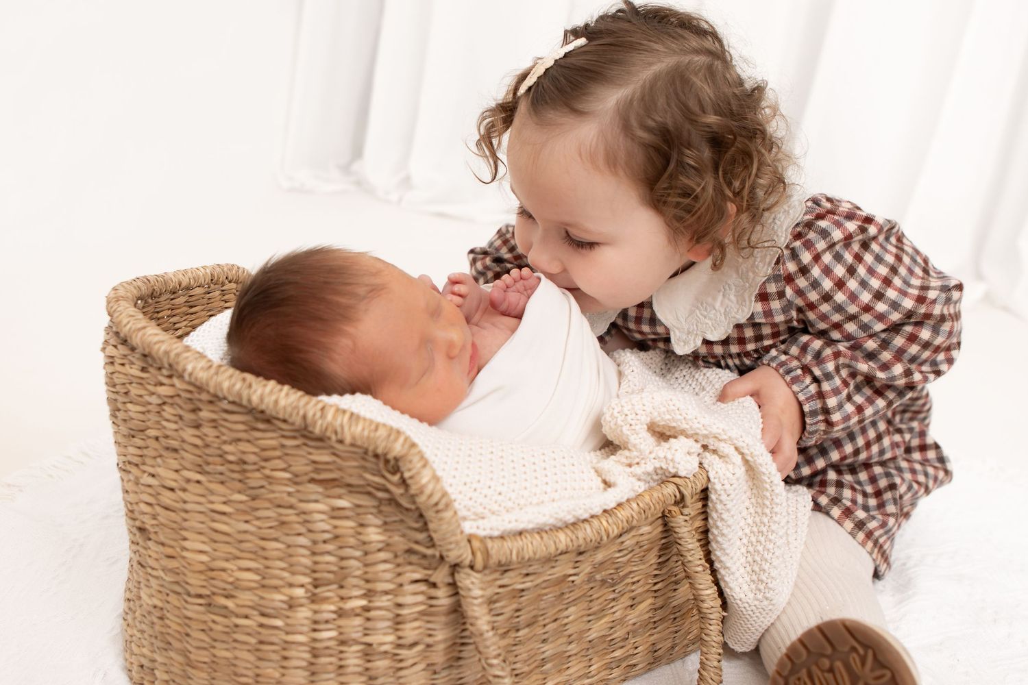 Toddler in checkered dress peeks at sleeping baby in woven basket.