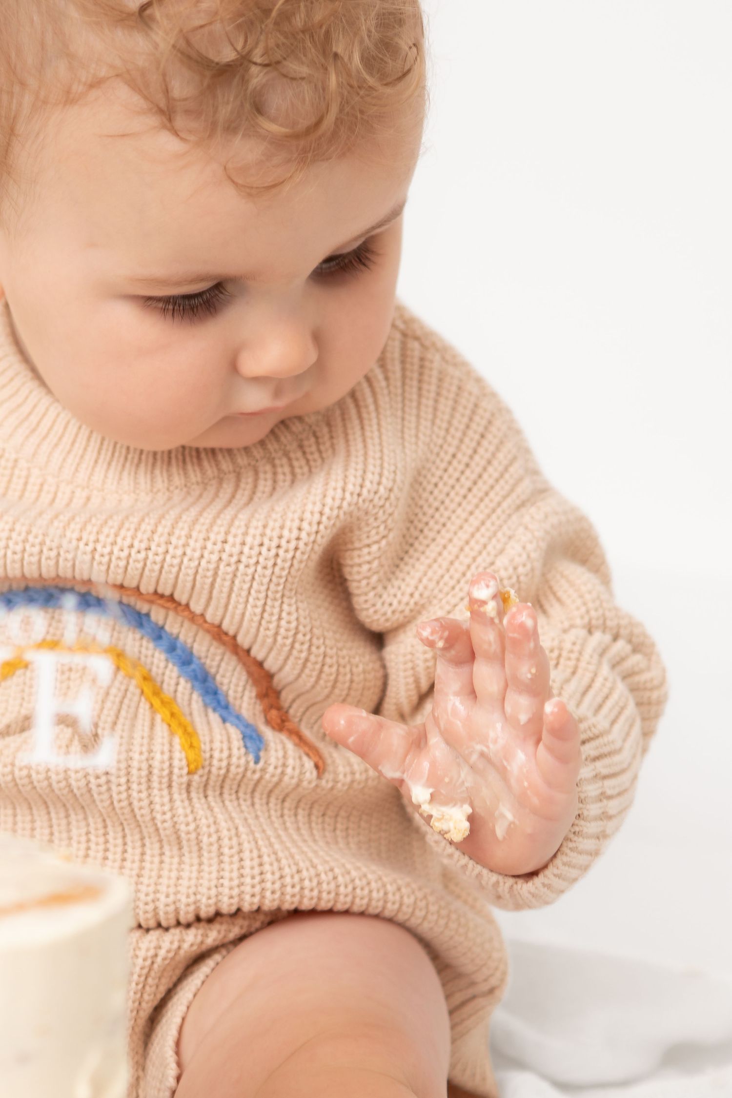 Cute baby in a beige rainbow knit sweater examining their frosting-covered hand, sitting against a white background.